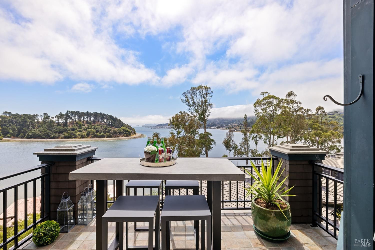 This image showcases a charming outdoor patio or balcony area with a stunning waterfront view. The space is furnished with a modern table and chairs, perfect for enjoying meals or relaxing outdoors. A potted plant adds a touch of greenery, while the railing provides safety without obstructing the scenic vista of the water and distant landscape.