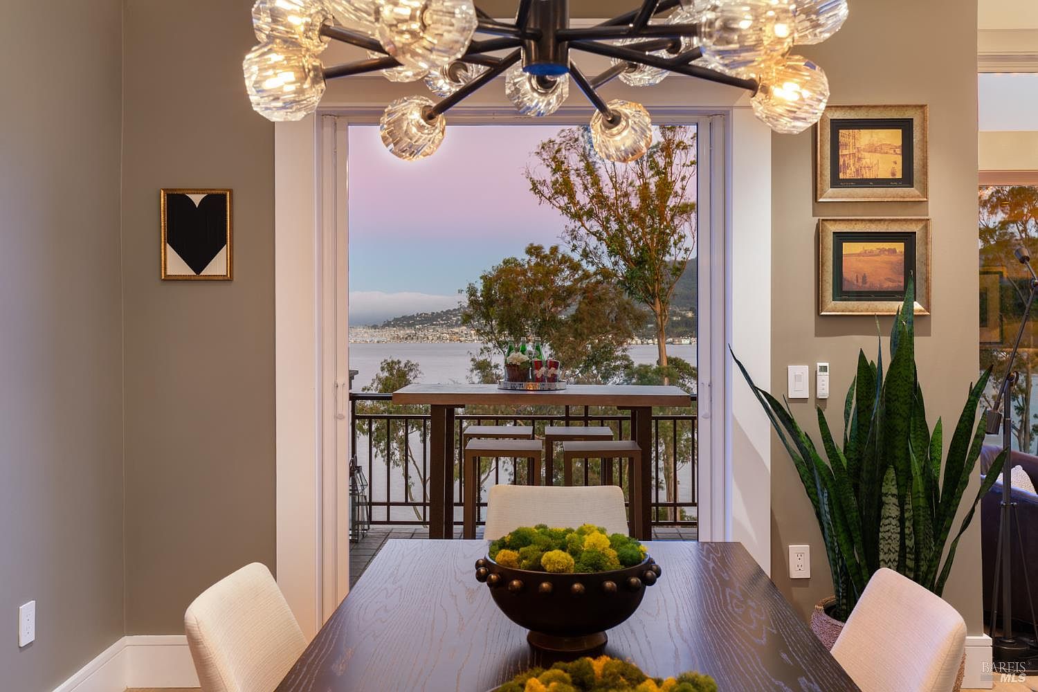 This interior shot showcases a dining room with a modern chandelier and a view to the outside. A dark wood dining table is centered, adorned with a decorative bowl, and surrounded by chairs. The open doorway leads to a balcony with a table and chairs, offering a scenic view of the water and distant cityscape.