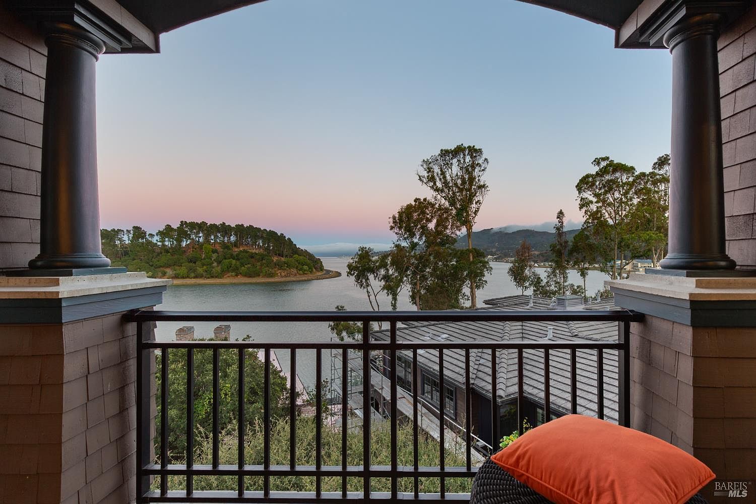 This image showcases a scenic view from a balcony, framed by dark wooden pillars and a black iron railing. The vista includes a tranquil body of water, a tree-covered island, and a glimpse of a neighboring house with a gray roof. An orange pillow on a wicker chair adds a touch of warmth and comfort to the outdoor space.