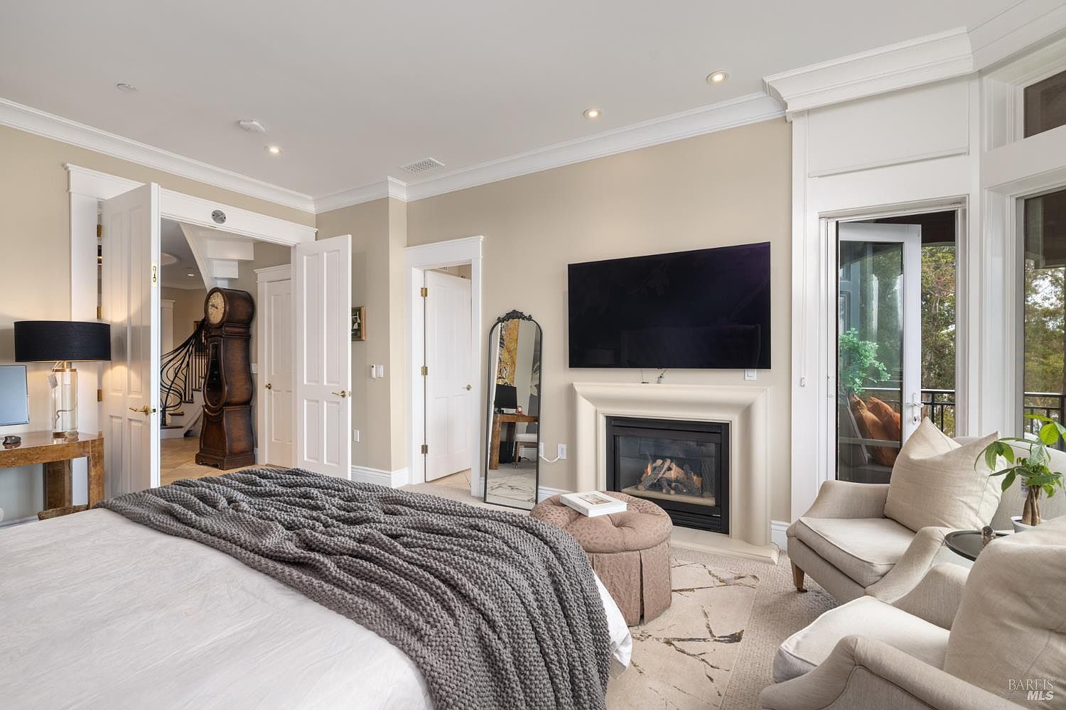 This is a primary bedroom featuring a king-size bed with a gray throw blanket, a fireplace with a mounted television above, and two armchairs near a balcony. The room has neutral-toned walls, white trim, and multiple doorways, including one leading to a staircase, and the overall style is luxurious and comfortable. The perspective is from the foot of the bed, looking towards the fireplace and balcony.