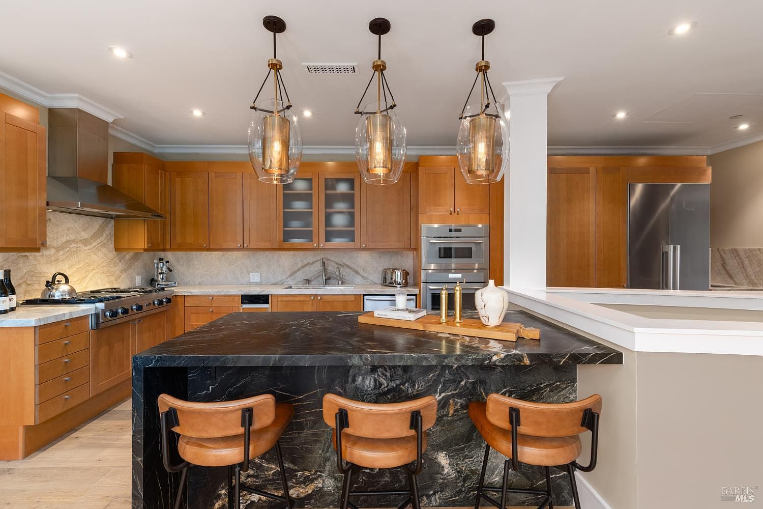 This is a well-lit kitchen featuring wooden cabinets, stainless steel appliances, and a dark marble island with seating. Three pendant lights hang above the island, adding a touch of elegance. The kitchen has a modern and sophisticated feel, with a focus on high-end finishes and functionality.