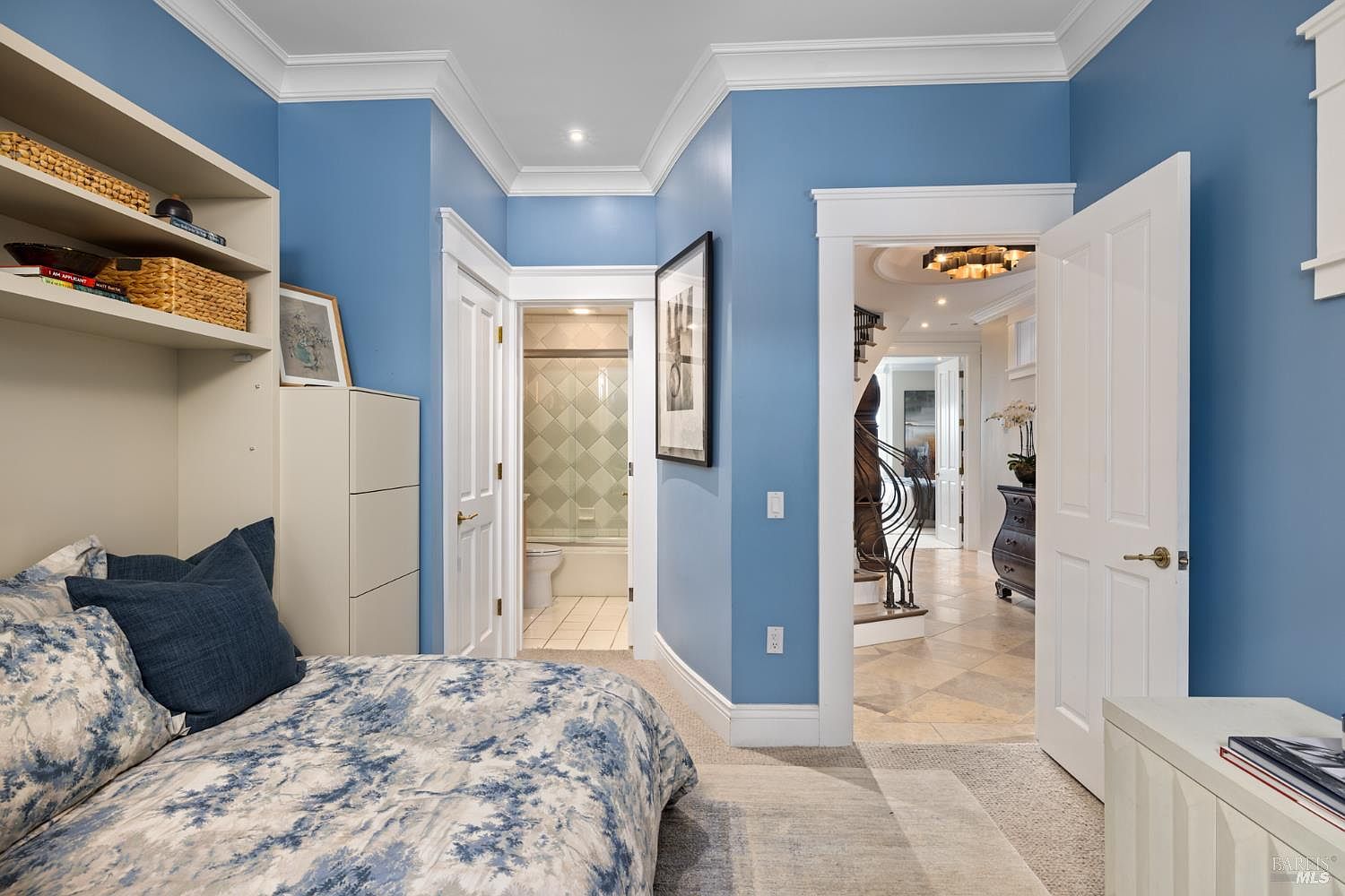This is a cozy guest bedroom featuring a built-in shelving unit and a bed with blue and white patterned bedding. The walls are painted a calming blue, complemented by white trim and doors. An open doorway leads to a hallway, while another door provides access to a bathroom, creating a convenient and private space for guests.