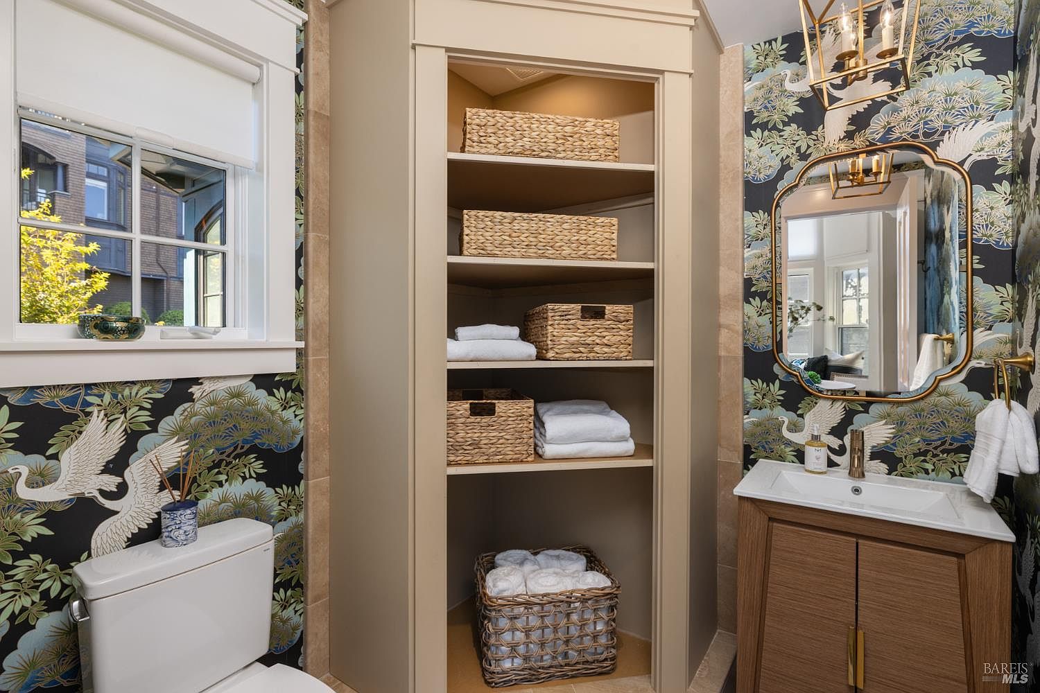 This is a well-decorated bathroom featuring a built-in linen closet with woven baskets and neatly folded towels. The vanity has a wood finish with a white countertop and a decorative gold-framed mirror. The wallpaper is a dark floral pattern with crane motifs, adding a touch of elegance and sophistication to the space.