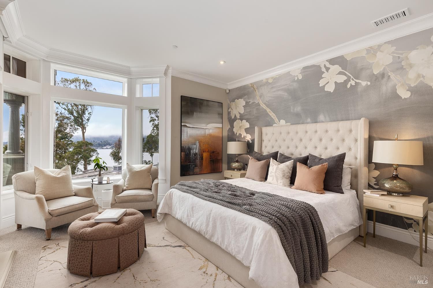 This is a luxurious primary bedroom featuring a large bed with a tufted headboard, complemented by a gray knit throw and decorative pillows. The room boasts a serene ambiance with a floral-patterned accent wall, a large abstract painting, and a bay window offering a scenic view of the lake. Two comfortable armchairs and an ottoman create a cozy seating area near the window.