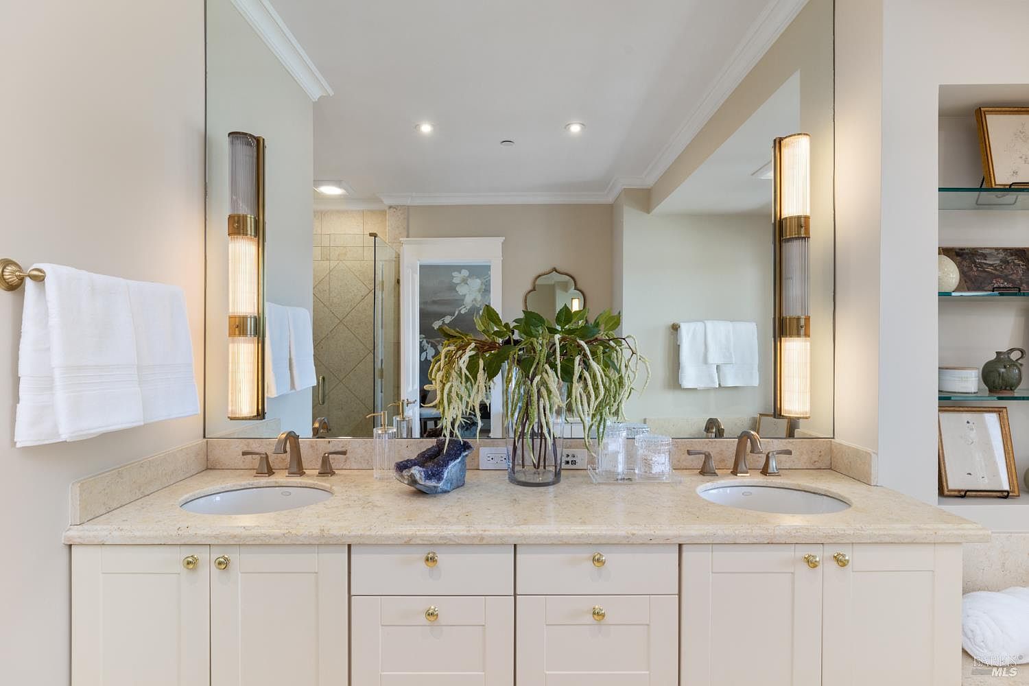 This is a well-lit primary bathroom featuring a double vanity with a light-colored countertop and cabinetry. The large mirror extends across the vanity, reflecting the lighting fixtures and creating a spacious feel. Decorative elements such as plants and artwork add a touch of elegance to the space.