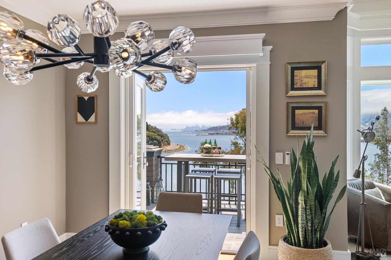 This dining room features a modern chandelier, a dark wood dining table with seating, and artwork on the walls. A large open doorway leads to a balcony with views of the water and city skyline. A potted snake plant adds a touch of greenery to the space.