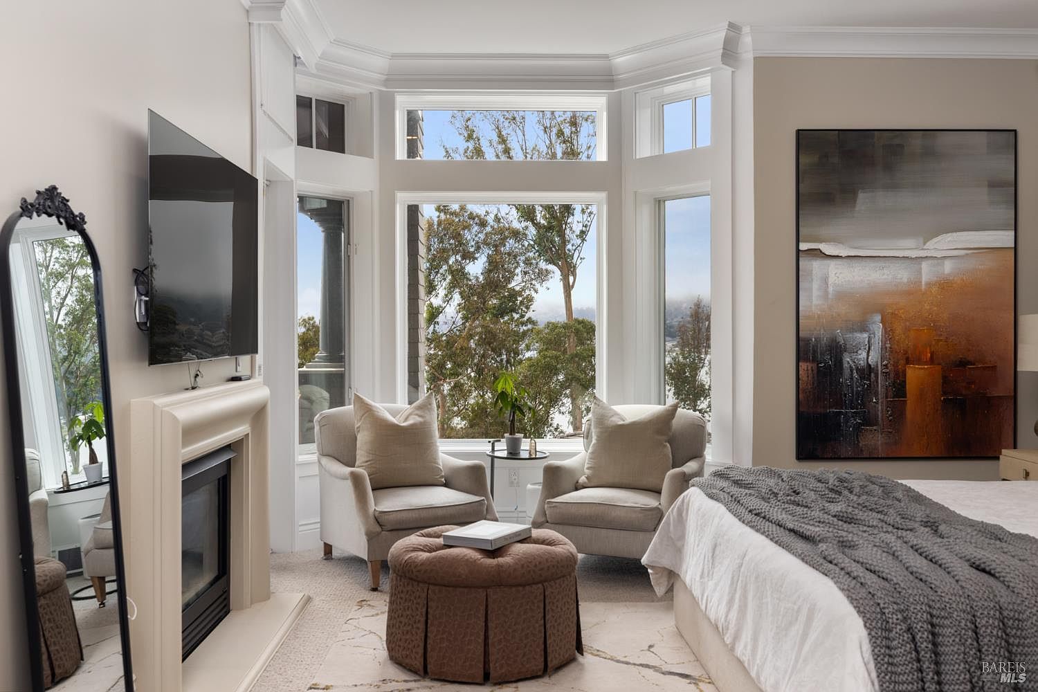 This is a primary bedroom featuring a sitting area with two armchairs and an ottoman in front of a large bay window. The room has a fireplace with a mounted television above it, and a large abstract painting hangs on the wall next to the bed. The style is elegant and comfortable, with neutral tones and natural light creating a relaxing atmosphere.