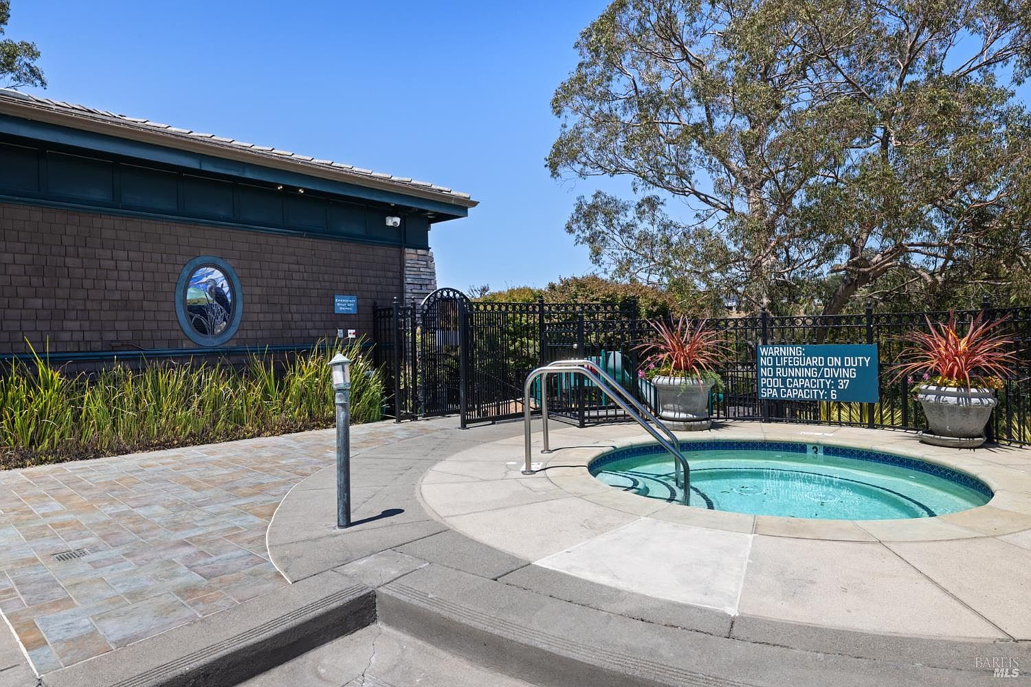 This image showcases a well-maintained spa area, featuring a circular hot tub with clear blue water, surrounded by a concrete deck and decorative planters. A black wrought iron fence encloses the area, providing privacy and safety. A building with a unique round window is visible in the background, adding architectural interest.