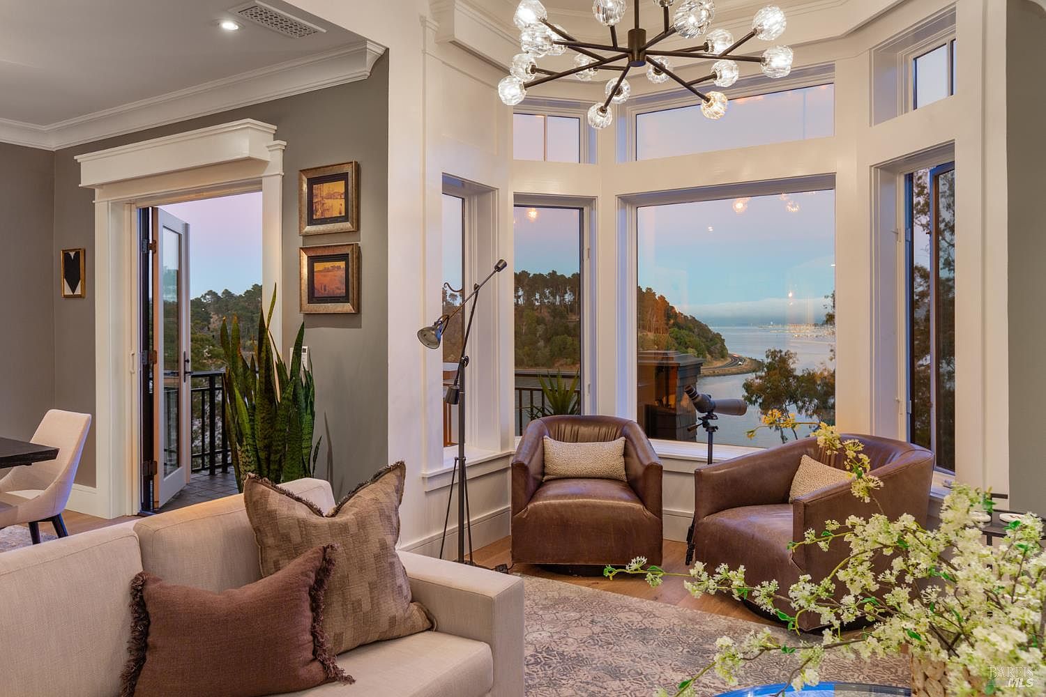 This is an interior shot of a luxurious living room featuring a bay window with a scenic view of the water and coastline. The room is furnished with comfortable leather armchairs, a neutral-toned sofa with decorative pillows, and a modern chandelier. The space is well-lit and exudes a sophisticated, inviting atmosphere.