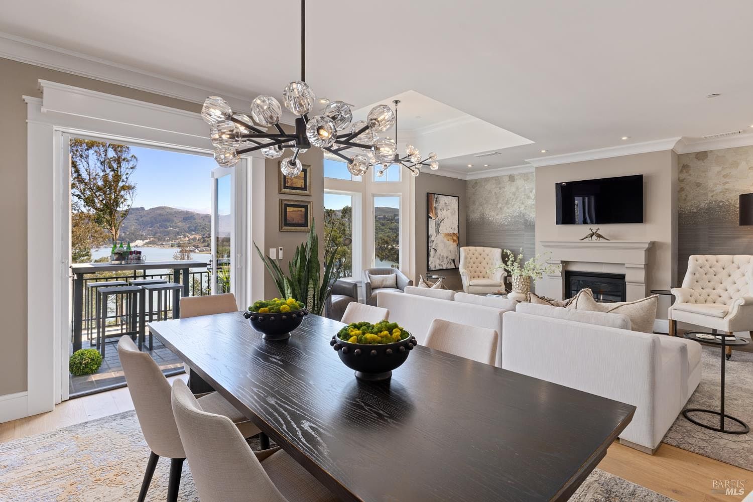 This is an interior shot of a dining room that seamlessly transitions into a living area, featuring a long, dark wood dining table with modern chairs. A striking chandelier hangs above the table, and large windows and an open balcony offer scenic views of the water and mountains. The living area includes a fireplace, a television, and comfortable seating, creating an inviting and luxurious atmosphere.