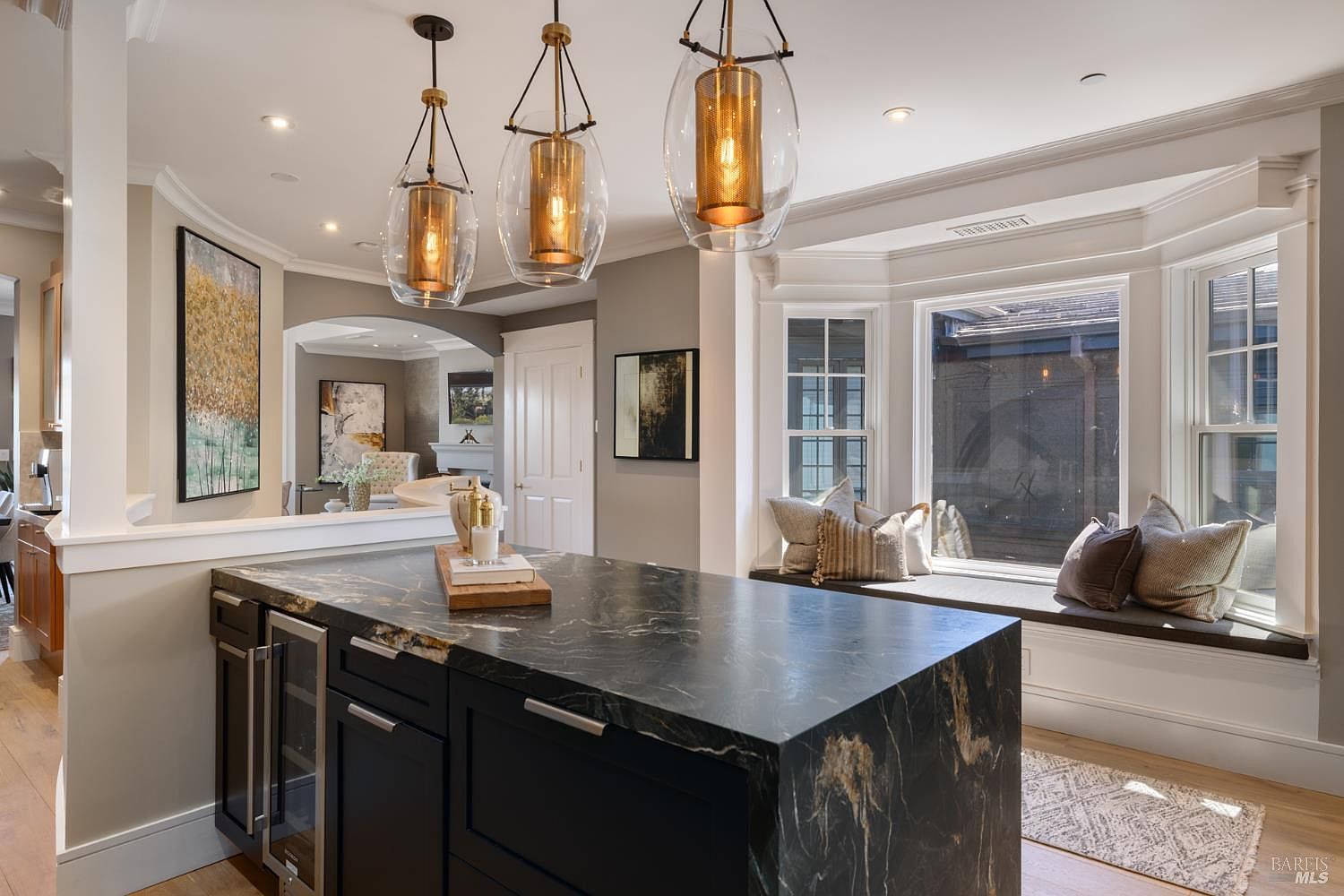 This is a well-lit kitchen featuring a dark marble island with a built-in wine cooler and modern pendant lighting. A window seat with decorative pillows is situated next to a bay window, offering a cozy nook. The kitchen opens to a living area, creating an open-concept space perfect for entertaining.