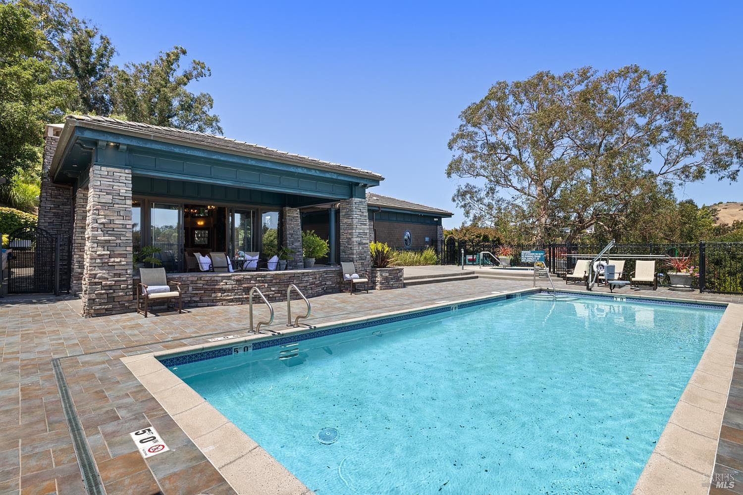 This image showcases a beautiful outdoor pool area, featuring a clear blue pool surrounded by a tiled deck. A covered lounge area with stone pillars and comfortable seating is adjacent to the pool, offering a relaxing space. Lounge chairs are placed near the pool, and the area is enclosed by a fence, providing privacy and safety.