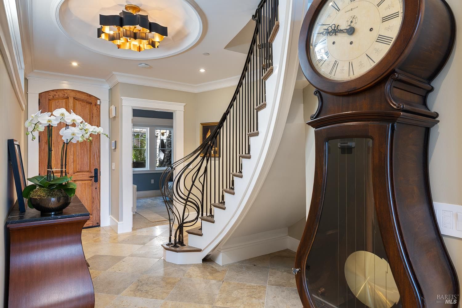 This interior shot showcases an elegant hallway with a curved staircase featuring wrought iron railings. A grandfather clock stands prominently to the right, while a wooden door and a console table with orchids add to the sophisticated ambiance. The flooring is tiled, and a decorative ceiling light fixture illuminates the space.