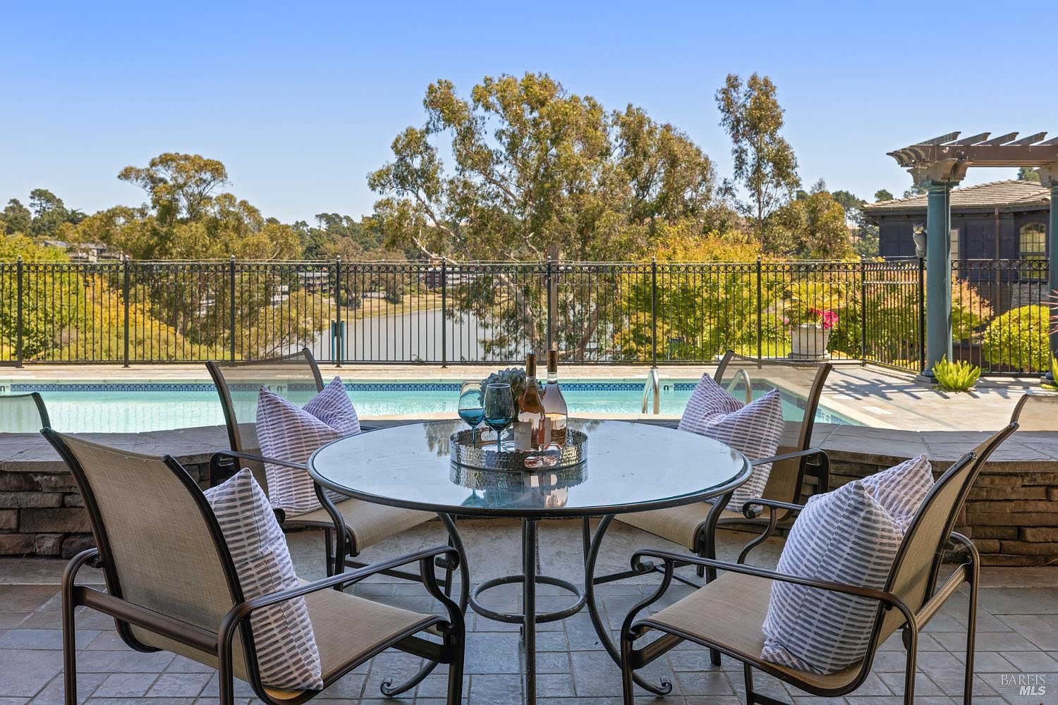 This image showcases a beautifully arranged outdoor patio area, complete with a round glass-topped table surrounded by comfortable chairs with striped cushions. The patio overlooks a sparkling pool and a scenic view of a lake and trees, creating an inviting and luxurious outdoor living space perfect for relaxation and entertaining.