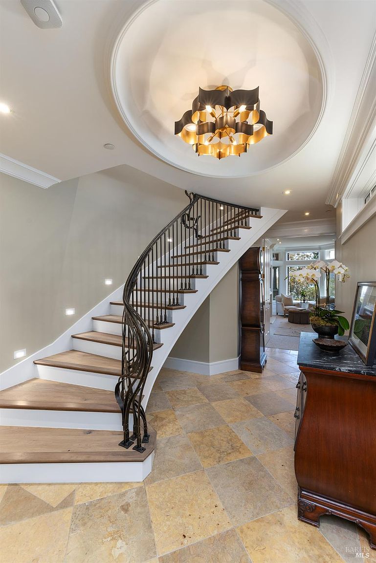 This interior shot showcases a grand foyer with a curved staircase, featuring wooden steps and an ornate iron railing. The travertine tile flooring adds warmth, while the statement chandelier and recessed lighting create an elegant ambiance. A glimpse into the living room suggests an open floor plan.
