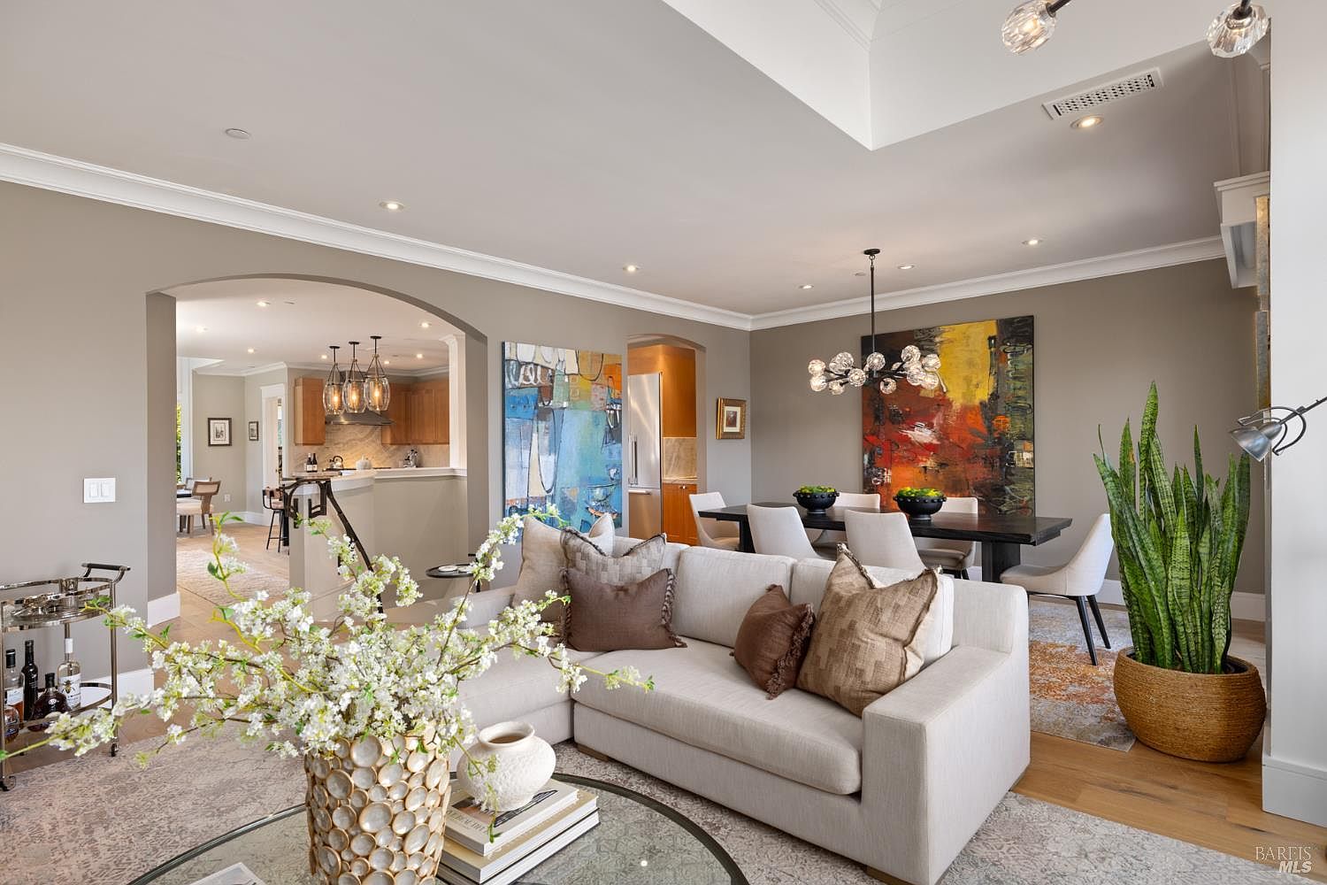 This is an interior shot of a living room and dining area. The living room features a sectional sofa with decorative pillows, a glass-topped coffee table with books and a vase of flowers, and a patterned area rug. The dining area includes a dark wood dining table with white chairs and a modern chandelier, with an abstract painting on the wall. A glimpse into the kitchen can be seen through an arched doorway.
