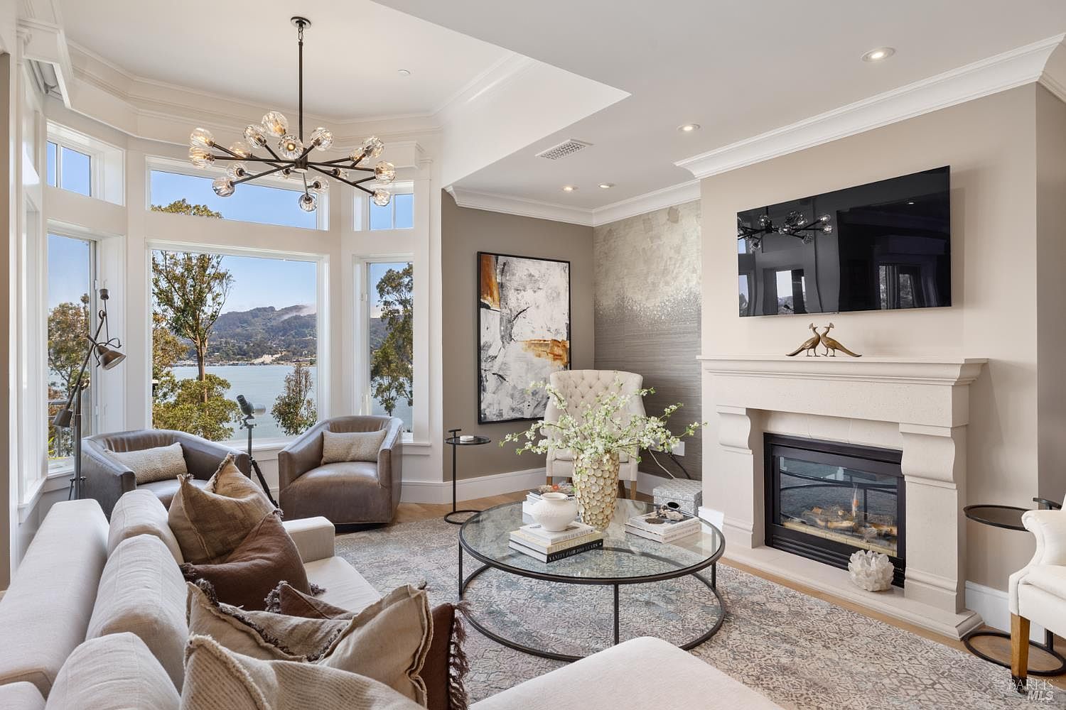 This is a beautifully staged living room featuring a neutral color palette, a large bay window with a water view, and a modern fireplace with a mounted television above. The room is furnished with comfortable seating, including a sectional sofa and armchairs, and a round glass coffee table adorned with decorative items. The overall impression is one of elegance and comfort, perfect for relaxation and entertaining.