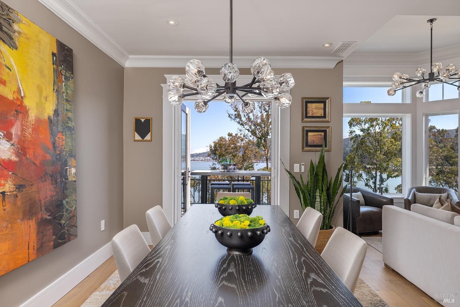 This is an interior shot of a dining room featuring a long, dark wood table centered under a modern chandelier. The room is decorated with contemporary art and opens to a balcony with a scenic water view. The space is well-lit and exudes a sophisticated, modern aesthetic.