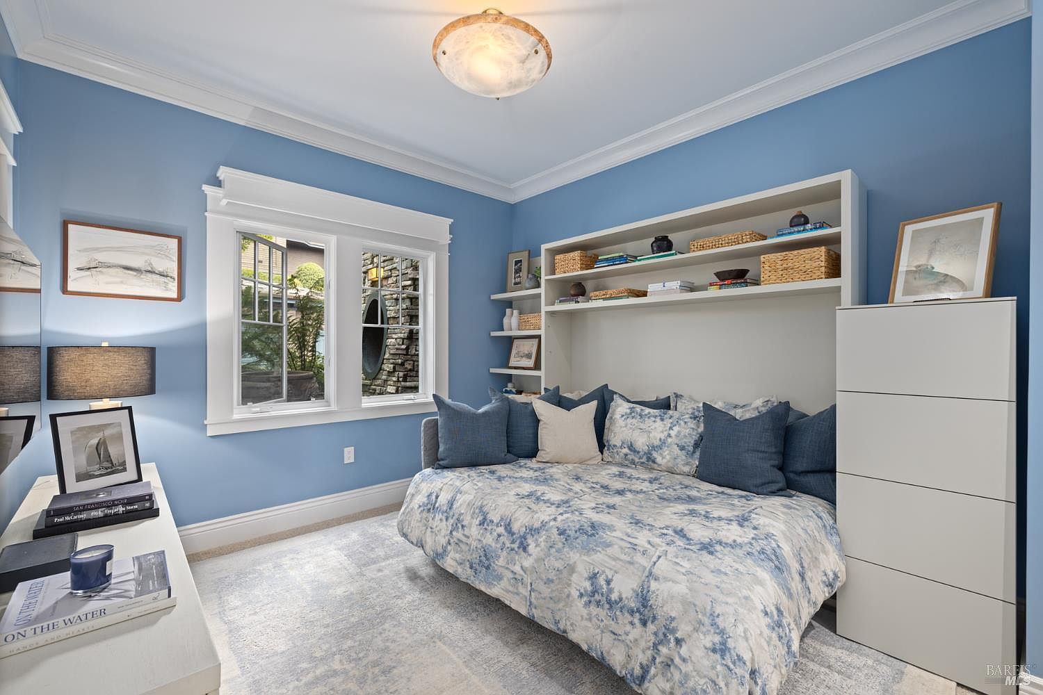 This is a cozy guest bedroom featuring blue walls and white trim, creating a serene atmosphere. The room includes a daybed with blue and white bedding, complemented by matching pillows. Built-in shelving and a dresser provide ample storage, while natural light streams in through a window, enhancing the room's inviting feel.