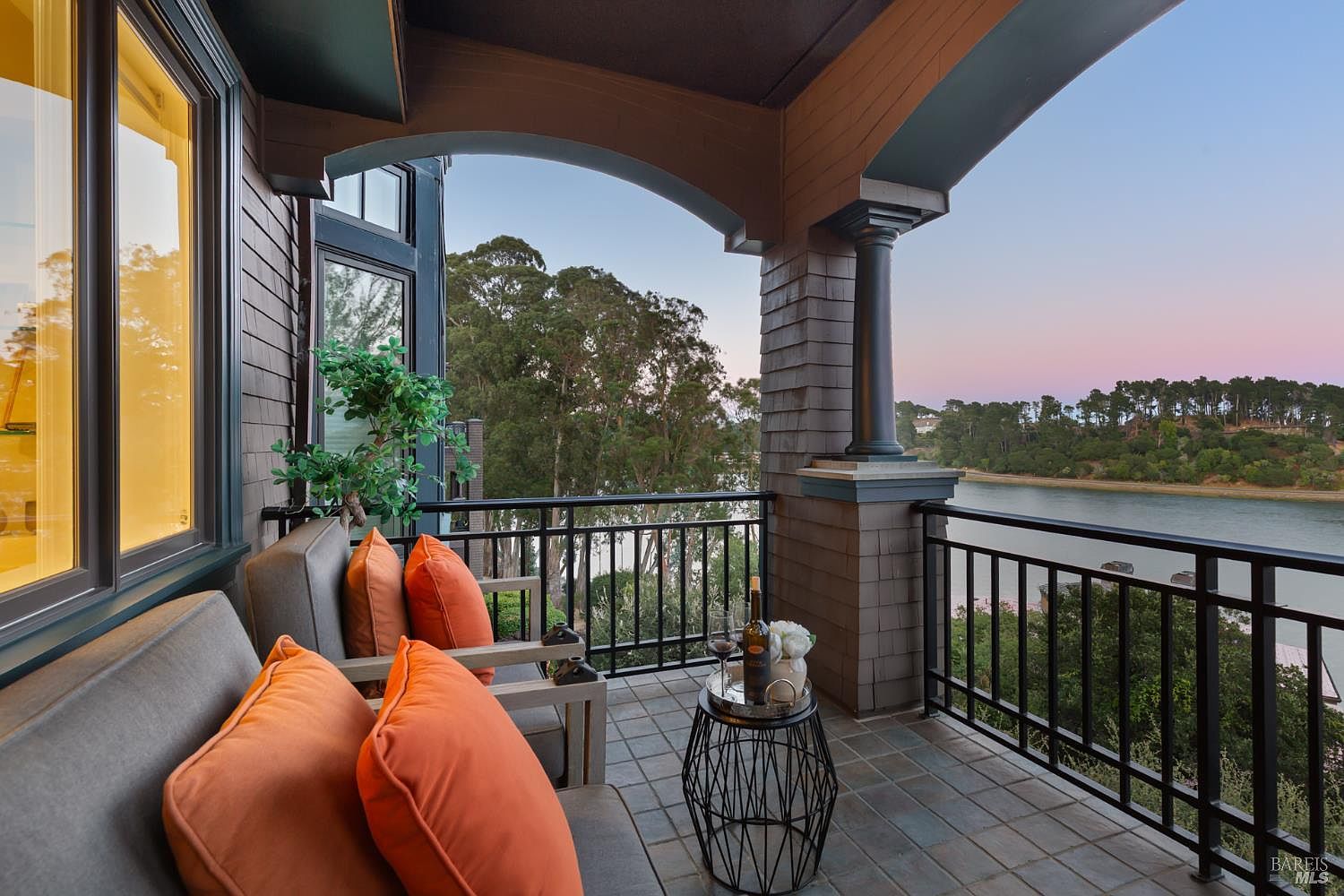 This image showcases a charming balcony with a serene waterfront view. The balcony features comfortable seating with orange pillows, a small table with wine and flowers, and a black railing. The architectural details include shingle siding and an arched opening, creating an inviting and relaxing outdoor space.