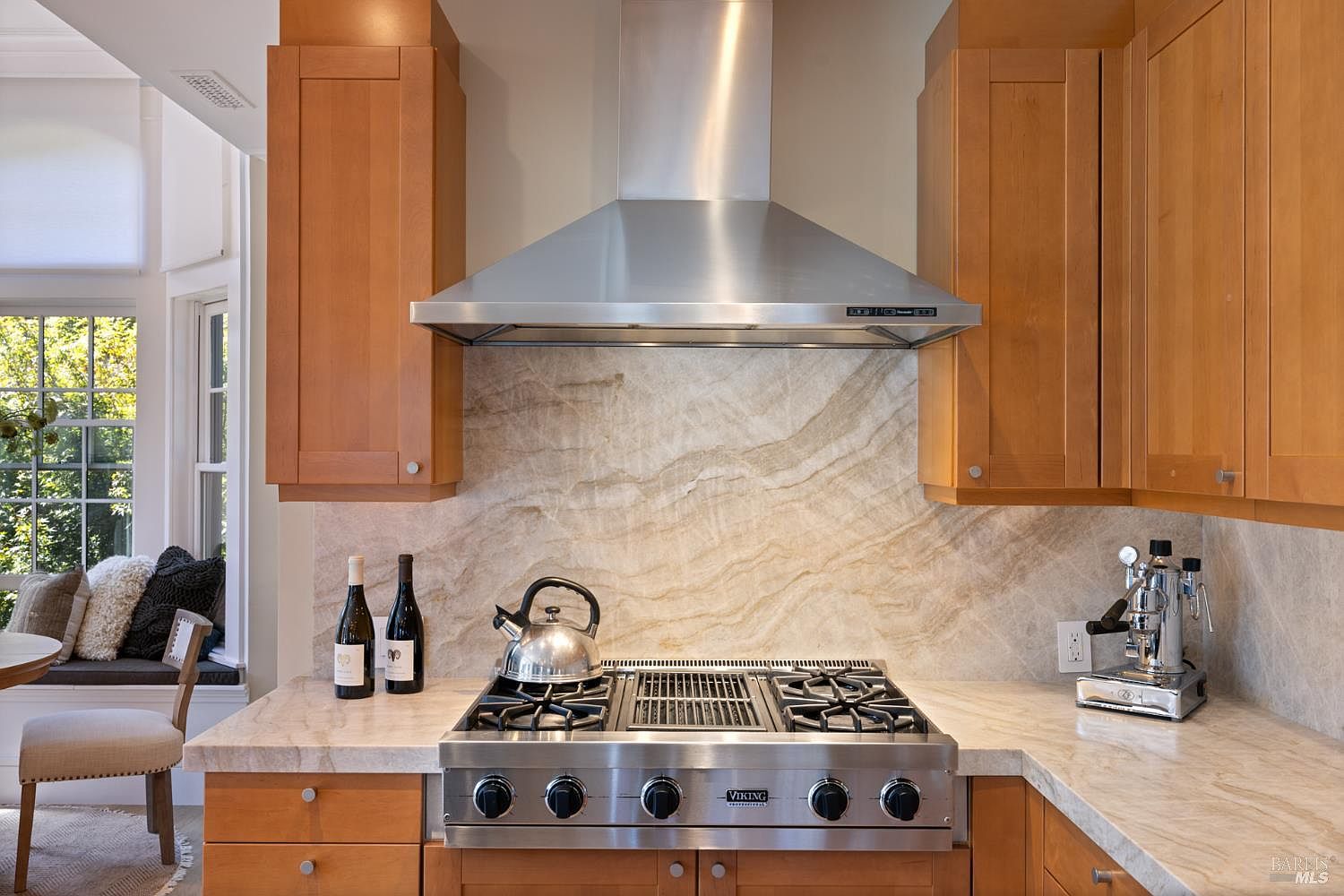 This is a well-appointed kitchen featuring wooden cabinetry, stainless steel appliances, and marble countertops. A Viking range and a stainless steel range hood are the focal points, complemented by a marble backsplash. The kitchen also includes a small seating area by a window, creating a cozy and inviting atmosphere.