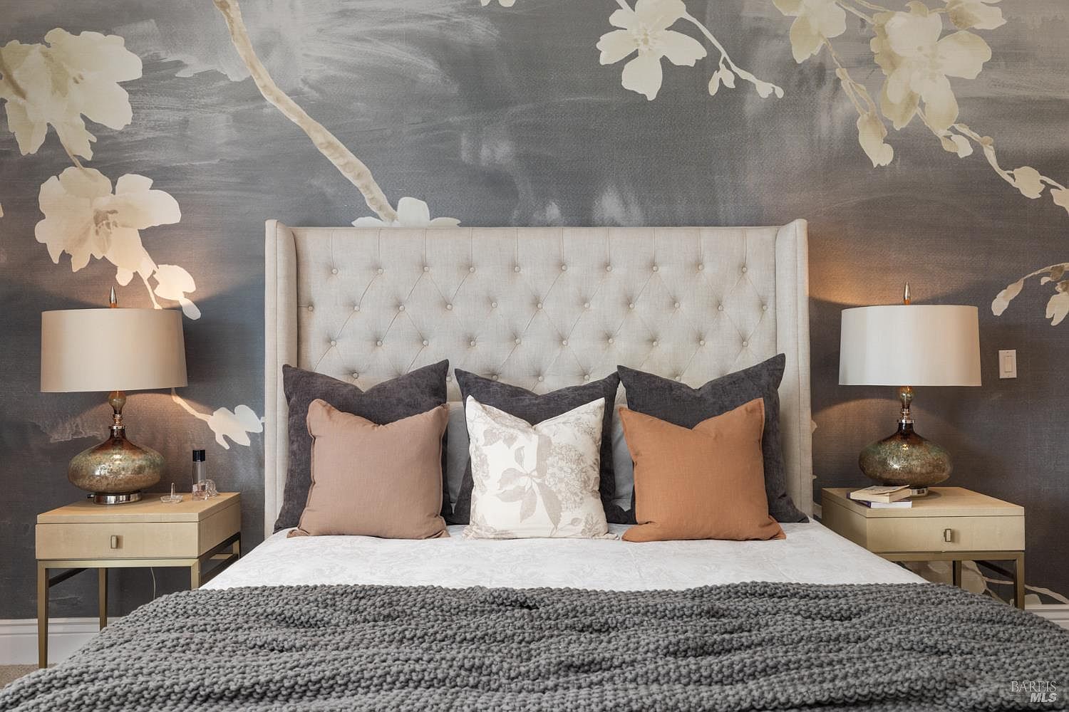 This is a beautifully staged primary bedroom featuring a tufted headboard, decorative pillows in neutral tones, and a cozy gray knit blanket. The room is illuminated by two elegant lamps on matching nightstands, and the wall is adorned with a subtle floral mural, creating a serene and inviting atmosphere. The perspective is a medium shot, focusing on the bed and headboard.