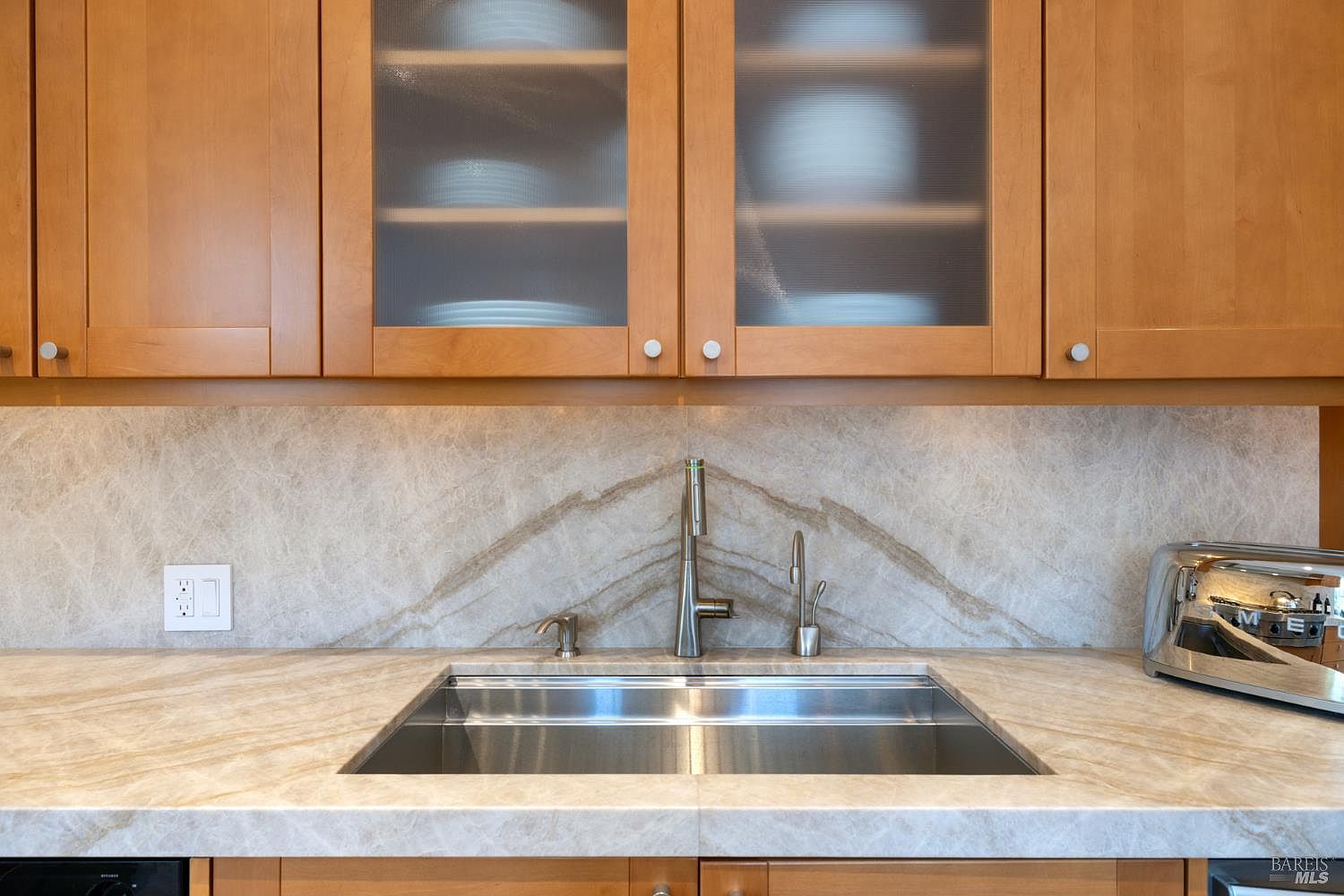 A well-lit kitchen showcases light wood cabinetry, some with frosted glass fronts, above a marble countertop and backsplash. A stainless steel sink is centered beneath a modern faucet, complemented by a matching soap dispenser and water filter tap. A stainless steel toaster sits on the counter to the right, adding a touch of modern convenience.