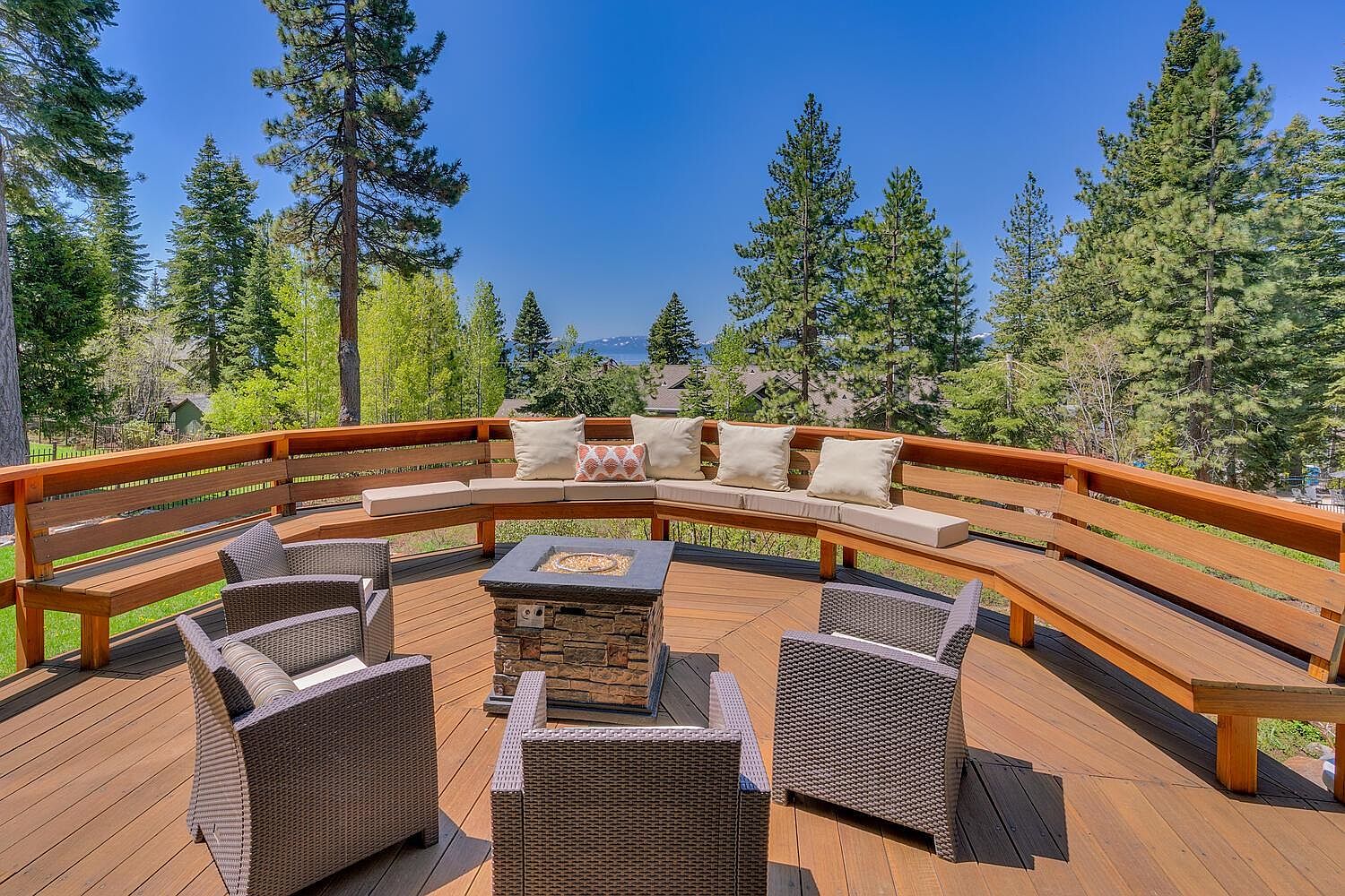 This image showcases a beautifully designed outdoor deck area, perfect for relaxation and entertainment. The deck features a built-in curved bench with comfortable cushions, surrounding a modern fire pit. Wicker chairs are arranged around the fire pit, creating an inviting seating arrangement, all set against a backdrop of lush green trees and a clear blue sky.