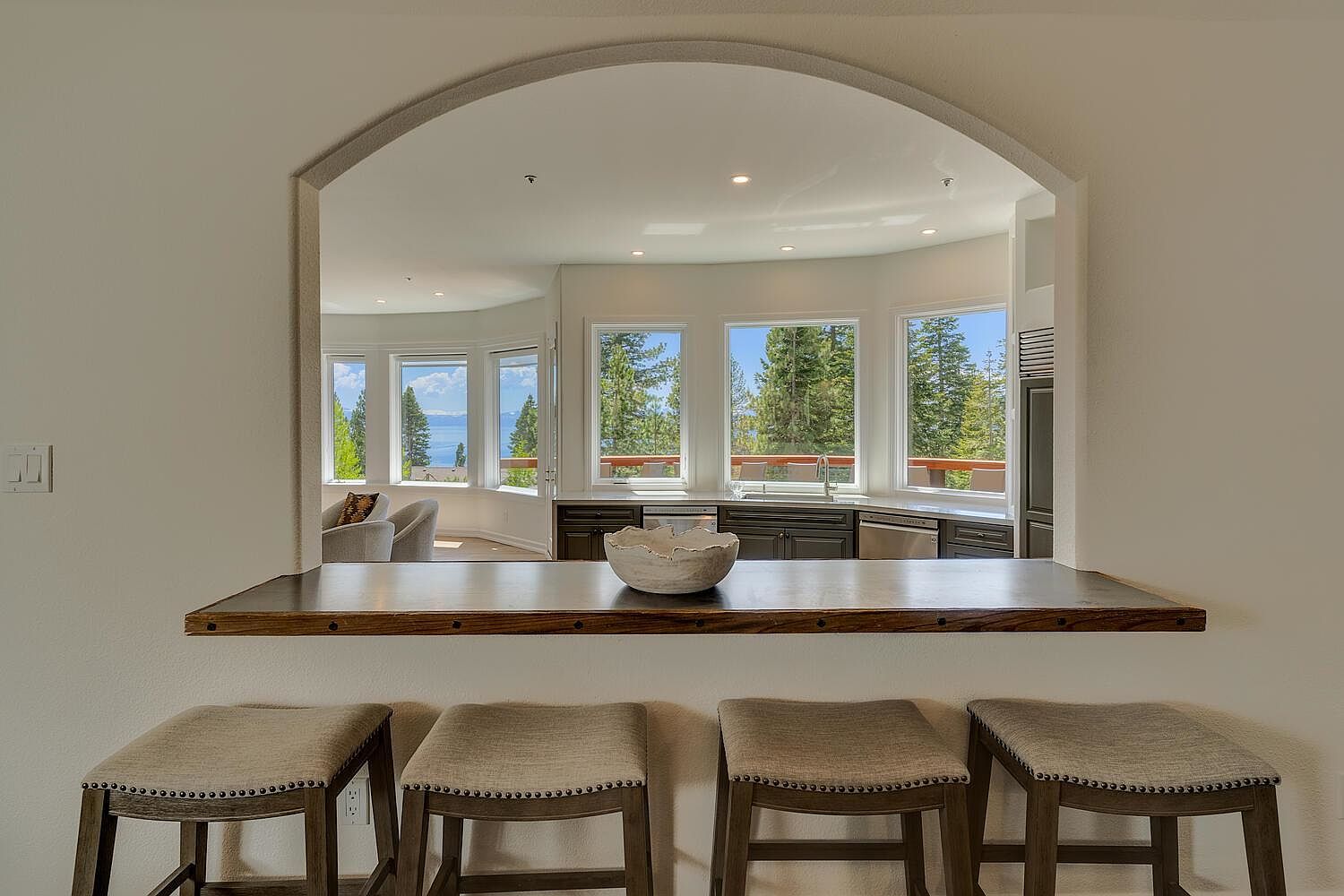 This interior shot showcases a modern kitchen with a unique arched opening that frames the view. The kitchen features dark cabinetry, stainless steel appliances, and a breakfast bar with four stools. The view through the windows reveals a scenic landscape with trees and a body of water, enhancing the kitchen's appeal.