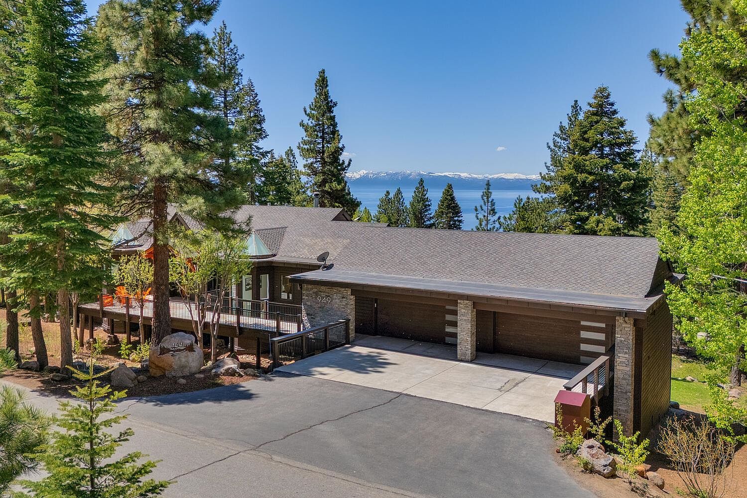 This is a front exterior view of a mountain home with a three-car garage. The house features a combination of wood and stone accents, a deck, and a paved driveway. In the background, there is a view of a lake and snow-capped mountains, enhancing the property's appeal.