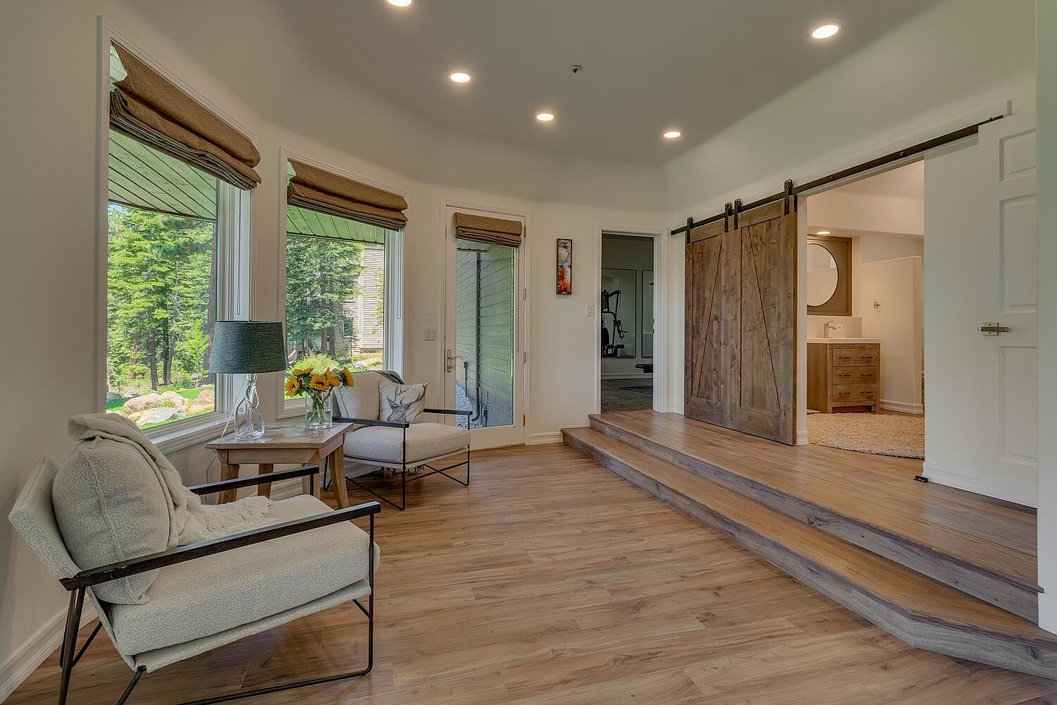 This interior shot showcases a cozy living room area with hardwood flooring and natural light streaming through large windows. The room features comfortable seating arrangements, including armchairs with neutral-toned cushions and a small side table with a lamp and floral arrangement. A unique barn door adds a rustic touch, leading to another room, while steps create a subtle elevation change within the space.