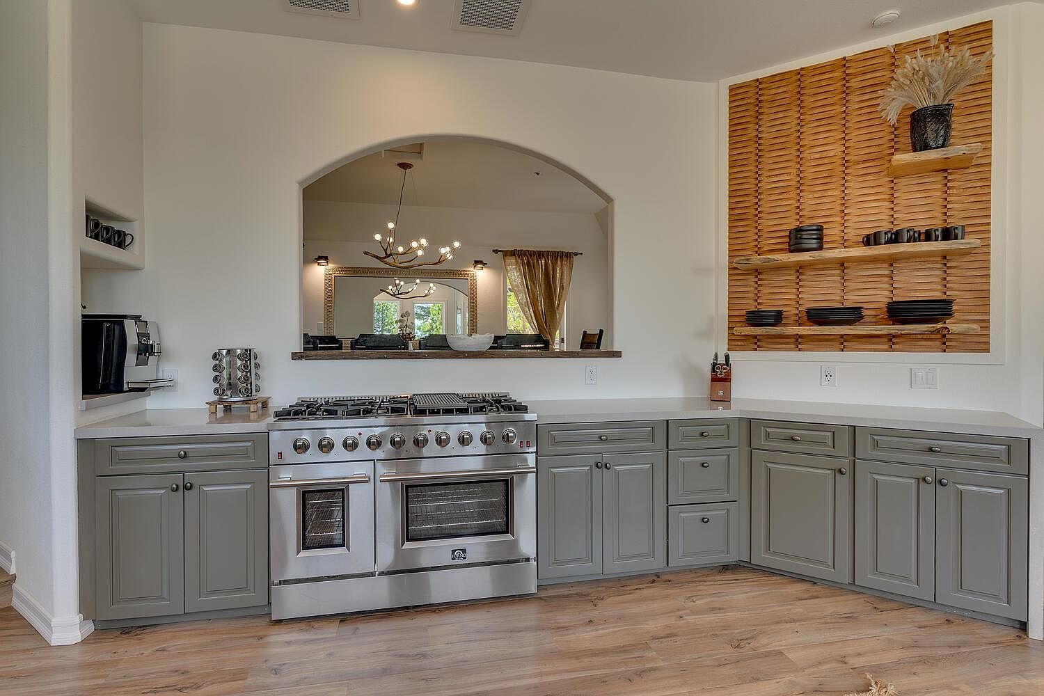 This is a well-lit kitchen featuring gray cabinets, stainless steel appliances, and light wood flooring. An arched opening provides a view into another room, while a decorative wooden panel with shelves adds visual interest. The kitchen has a clean and modern aesthetic.