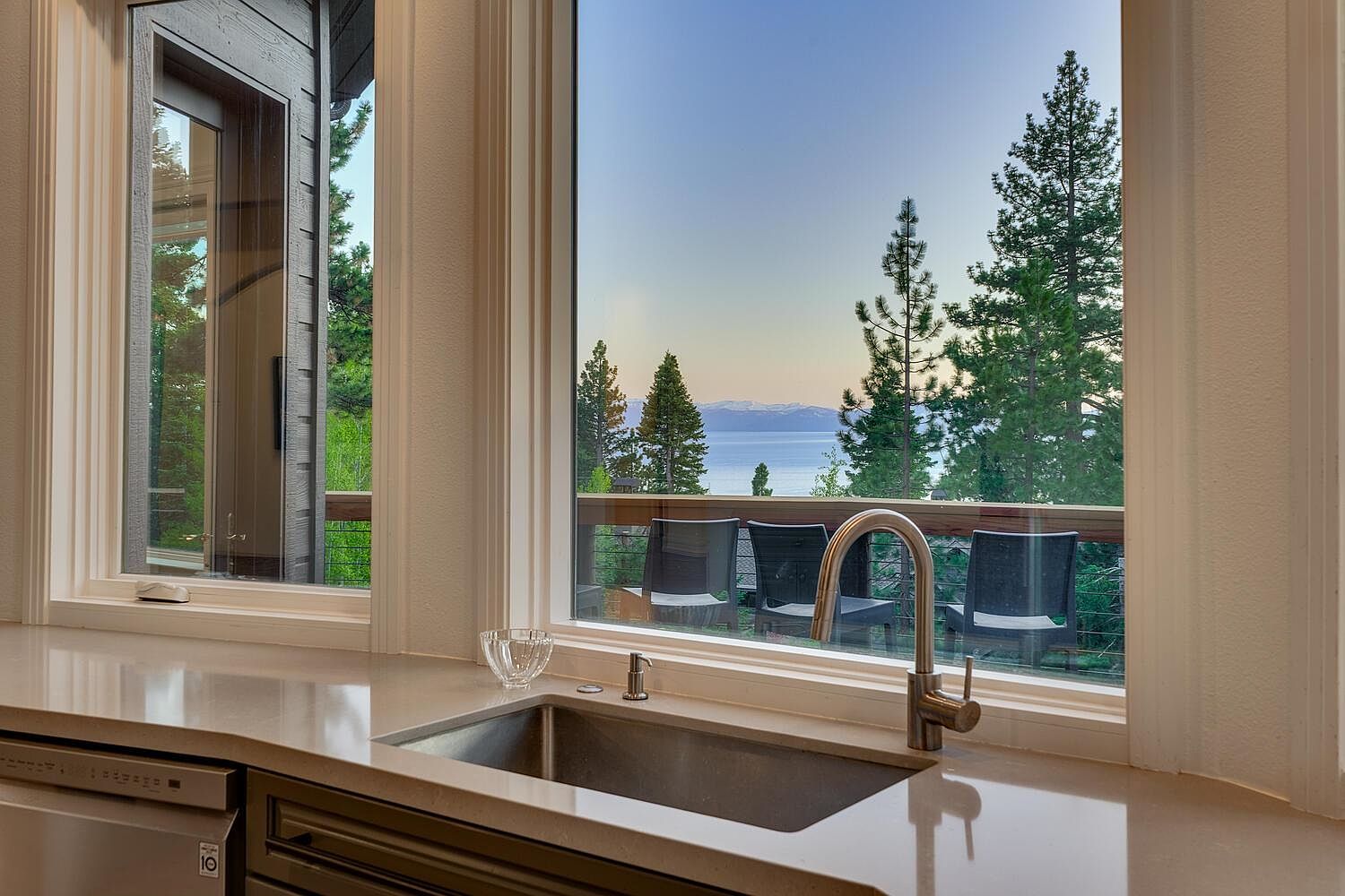 This image showcases a modern kitchen interior, focusing on the sink area with a large window providing a scenic view of a lake and trees. The countertop is a light, solid surface, and the sink is stainless steel with a modern faucet. The window offers a glimpse of an outdoor deck with chairs, enhancing the connection between indoor and outdoor living.