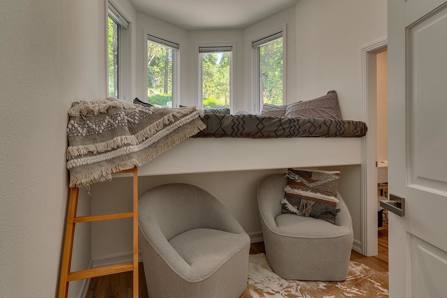 This interior shot showcases a cozy bedroom nook featuring a built-in bed platform with a patterned mattress and pillows, accessible by a wooden ladder. Below the bed, two rounded armchairs sit on a cowhide rug, creating a comfortable seating area. Natural light streams in through multiple windows, enhancing the room's inviting atmosphere.