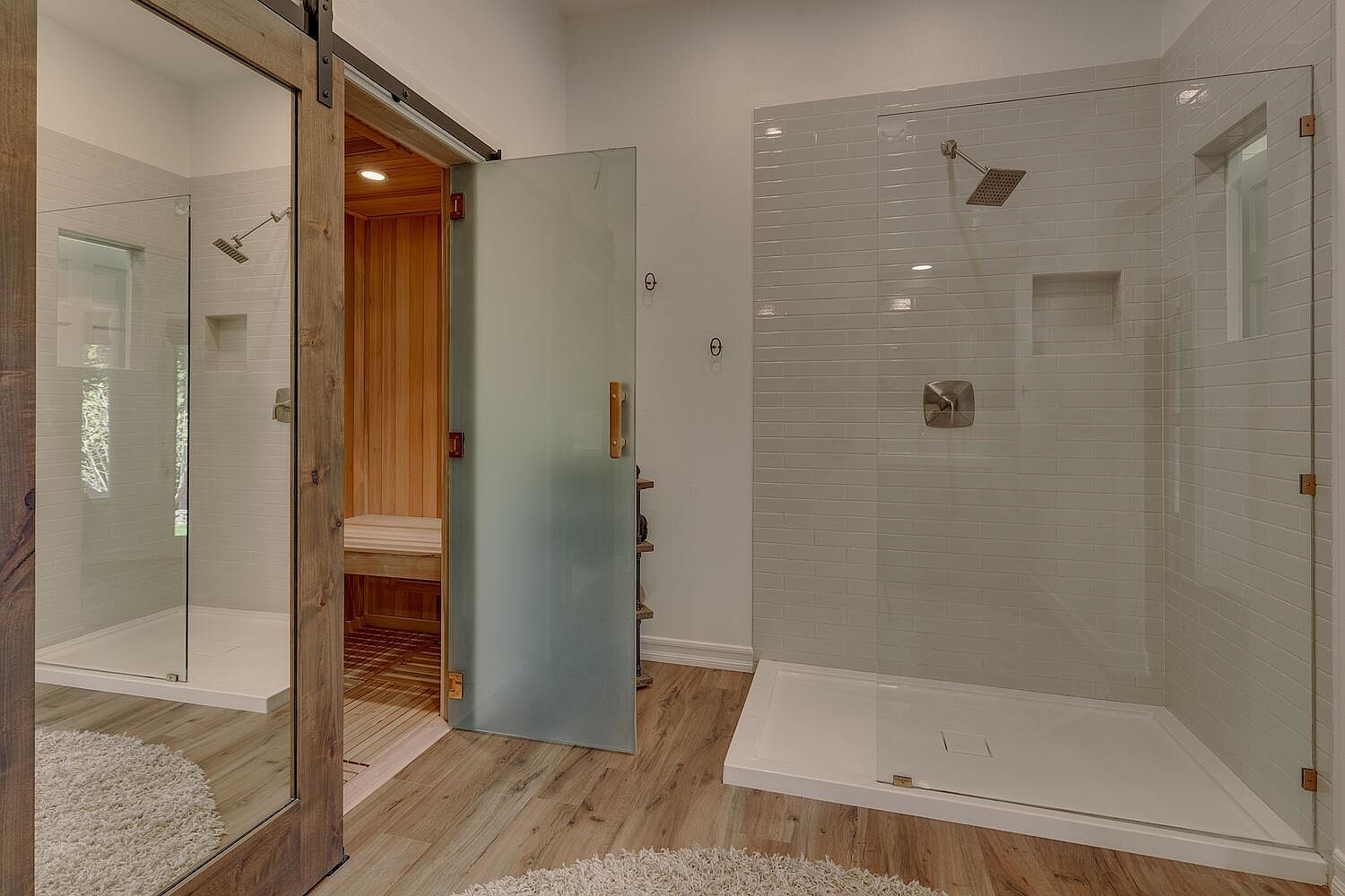 This is a luxurious primary bathroom featuring a modern shower with white subway tile and glass enclosure, complemented by a sauna with a frosted glass door. The bathroom has wood-look flooring and a neutral color palette, creating a spa-like atmosphere. The sliding barn door adds a rustic touch to the contemporary design.