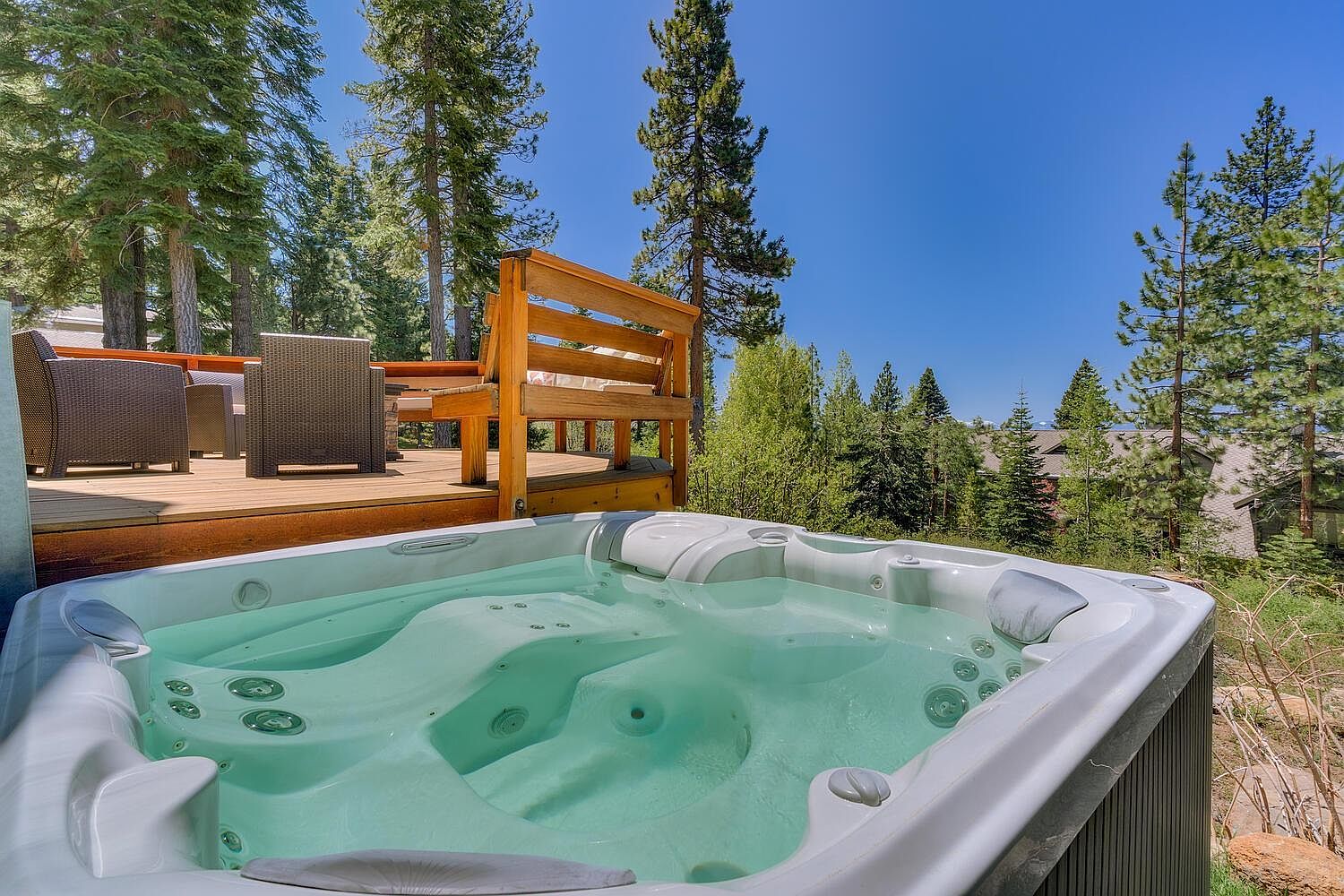 This image showcases a luxurious outdoor spa area, featuring a modern hot tub filled with clear, inviting water. The spa is surrounded by a wooden deck with outdoor seating and lush greenery, creating a serene and private retreat. Tall trees frame the scene, enhancing the sense of seclusion and natural beauty.