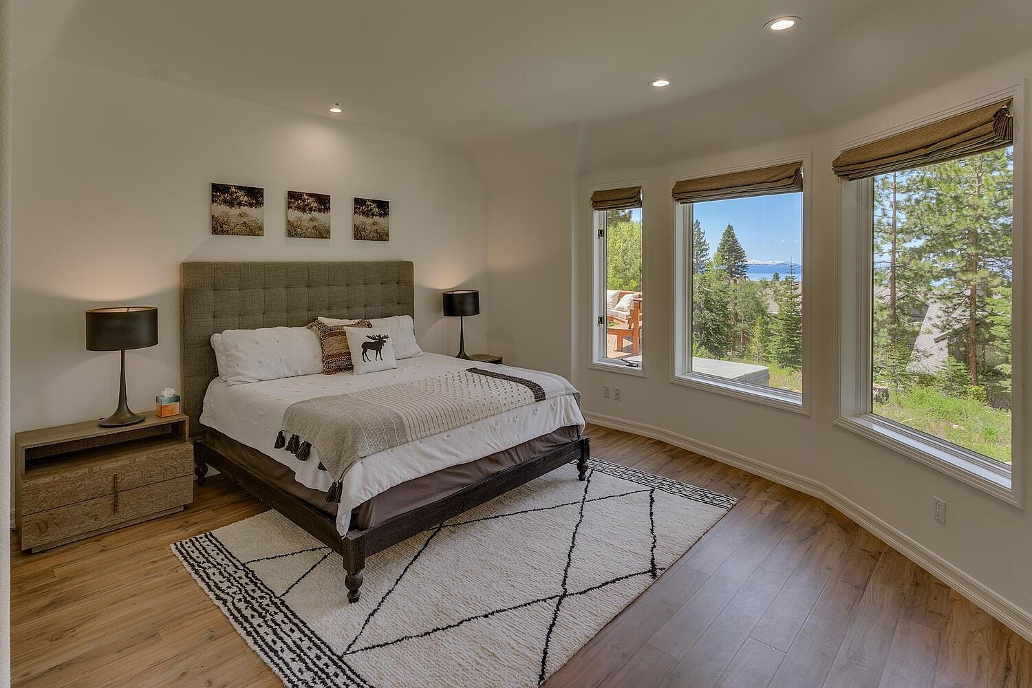 This is a primary bedroom featuring a large bed with a tufted headboard, flanked by nightstands and lamps. The room has a neutral color palette with white walls and a patterned area rug. Large windows offer a view of the surrounding landscape, bringing natural light into the space.
