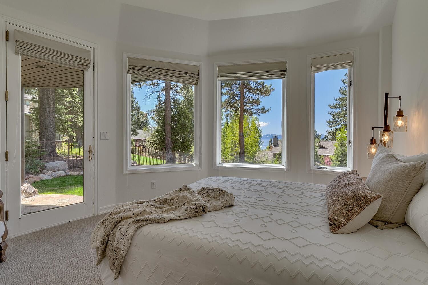 This is a bright and airy primary bedroom featuring a large bed with a patterned white comforter and decorative pillows. The room has multiple windows offering views of the surrounding trees and a glimpse of a lake. A glass door leads to an outdoor patio, enhancing the connection to nature.