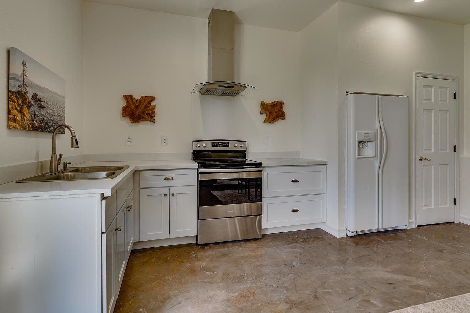 This is a well-lit kitchen featuring white cabinets, stainless steel appliances, and light countertops. The kitchen includes a sink, stove with a range hood, and a refrigerator. The floor has a unique, mottled pattern, and the overall impression is clean and functional.