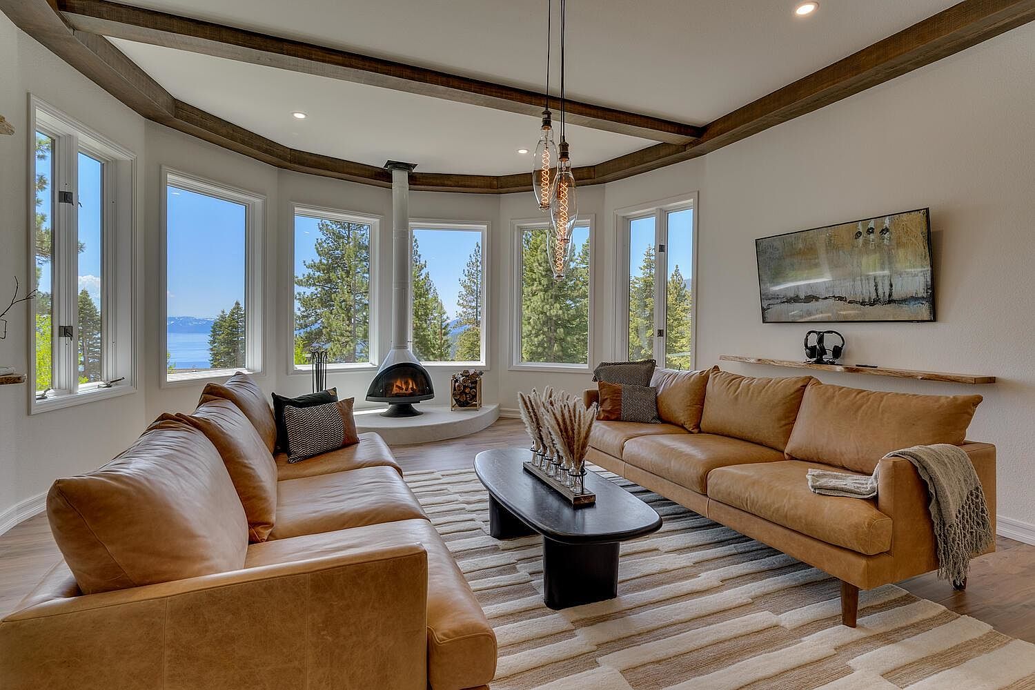This is an interior shot of a living room featuring two tan leather sofas facing each other on a striped rug. A modern fireplace sits in the corner surrounded by large windows with views of trees and a lake. The room has wooden beams on the ceiling and a modern black coffee table.