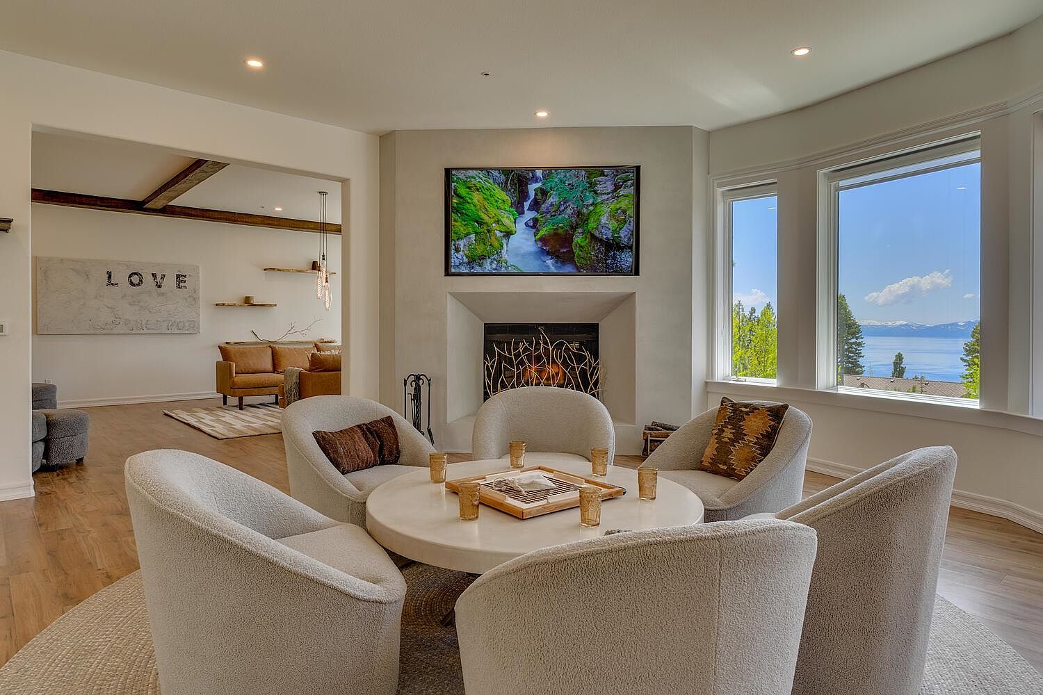 This is a cozy and inviting living room featuring a circular seating arrangement around a round table, perfect for conversation. A modern fireplace with a mounted television above it serves as a focal point, while large windows offer a scenic view of the outdoors. The room is decorated in neutral tones, creating a serene and comfortable atmosphere.