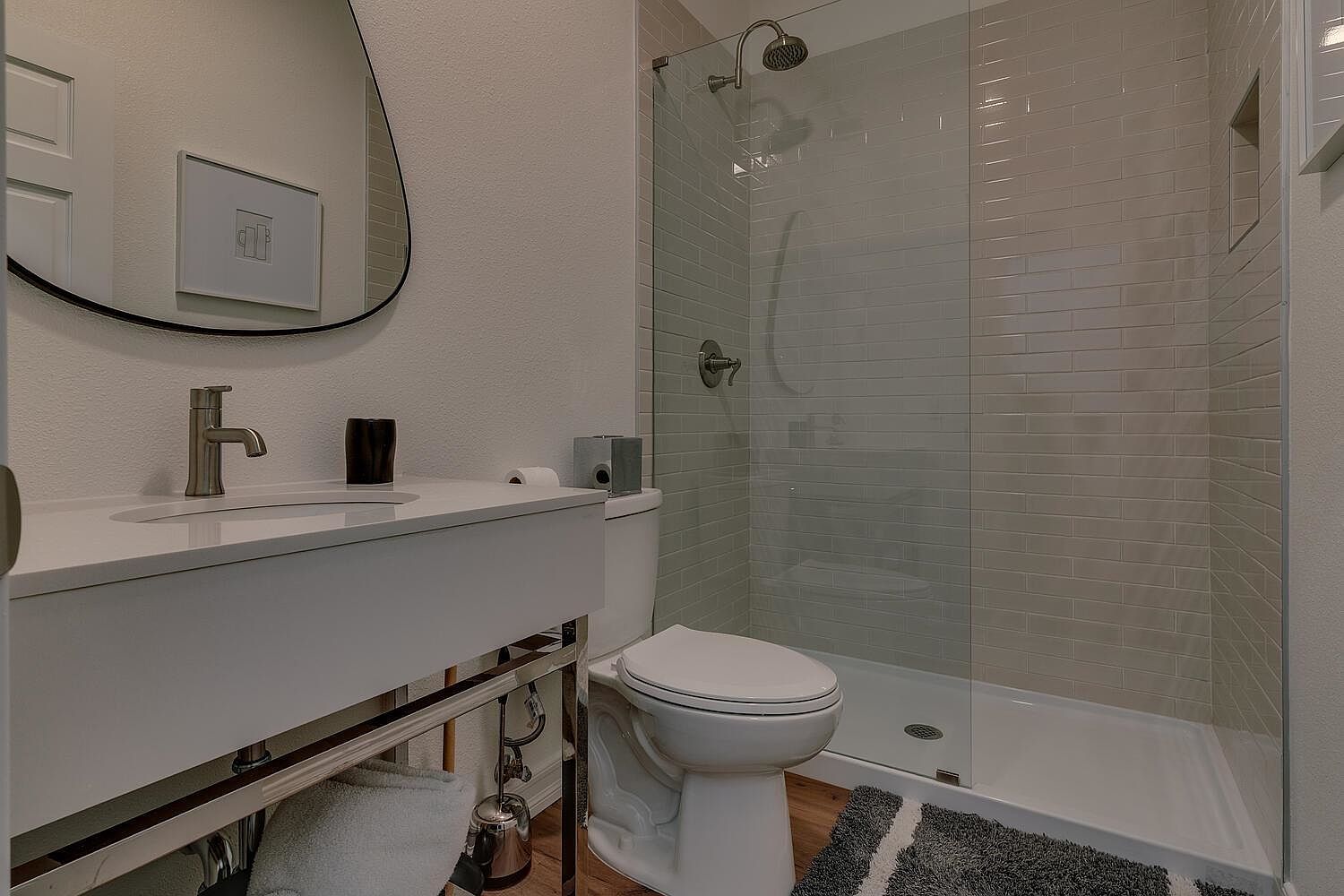 This is a well-lit guest bathroom featuring a modern vanity with a white countertop and chrome fixtures. A sleek, frameless shower enclosure with light gray subway tile adds a contemporary touch. The room is completed with a toilet and a stylish oval mirror, creating a clean and inviting space.