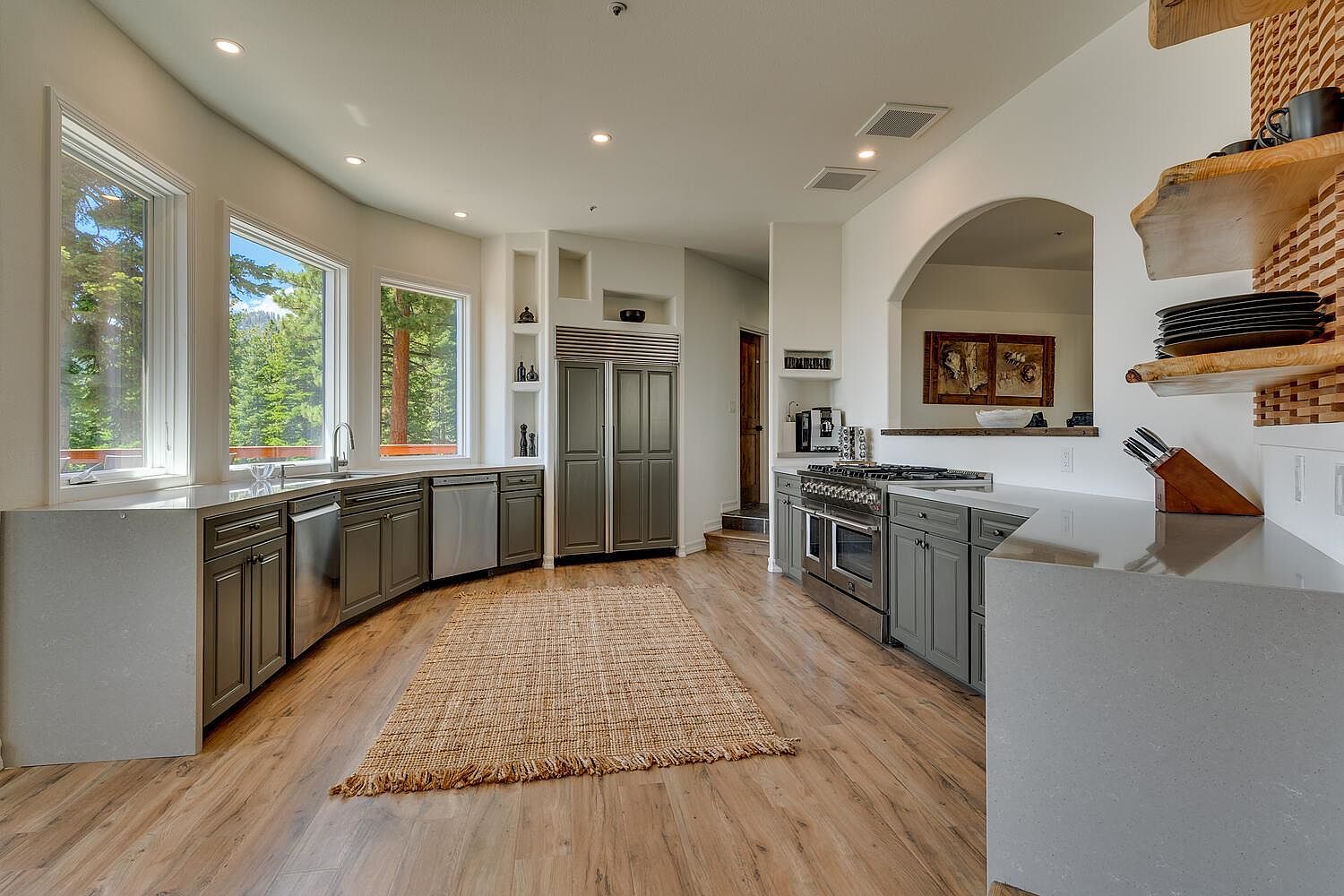 This is a spacious kitchen with a curved layout featuring gray cabinetry, stainless steel appliances, and light-colored countertops. Large windows offer views of the surrounding trees, and a woven rug adds texture to the hardwood floors. The kitchen also includes a built-in refrigerator and a pass-through to another room, creating an open and inviting atmosphere.