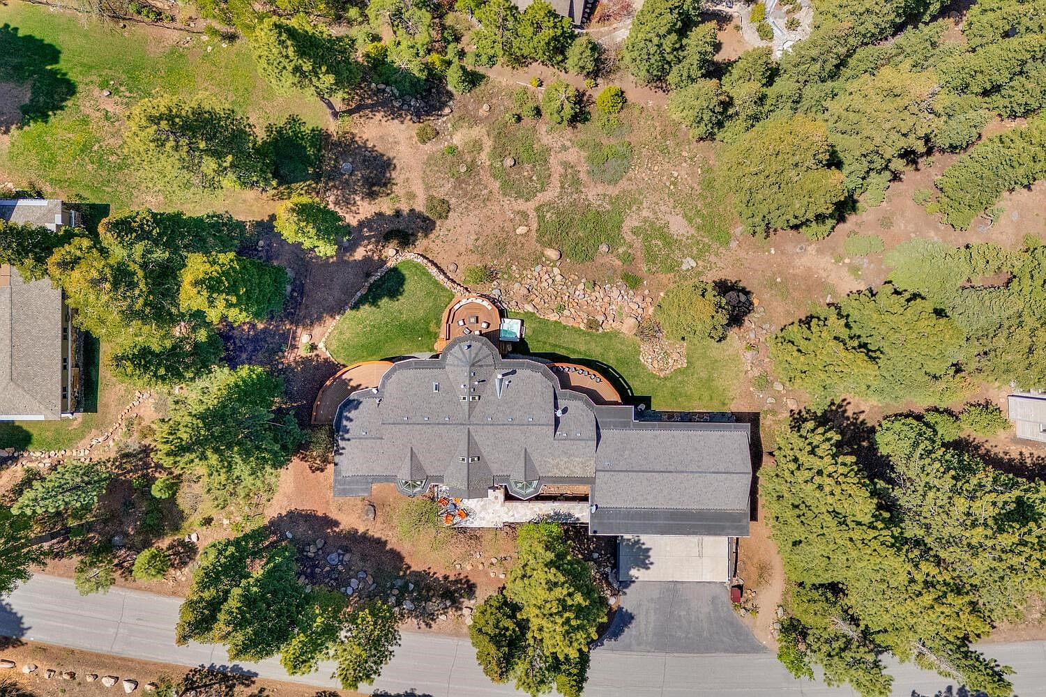 This aerial view showcases a luxurious home nestled among lush greenery. The house features a complex roofline, a spacious deck with outdoor seating and a hot tub, and a long driveway leading to a garage. The surrounding landscape includes mature trees, manicured lawns, and natural rock formations, creating a private and serene setting.