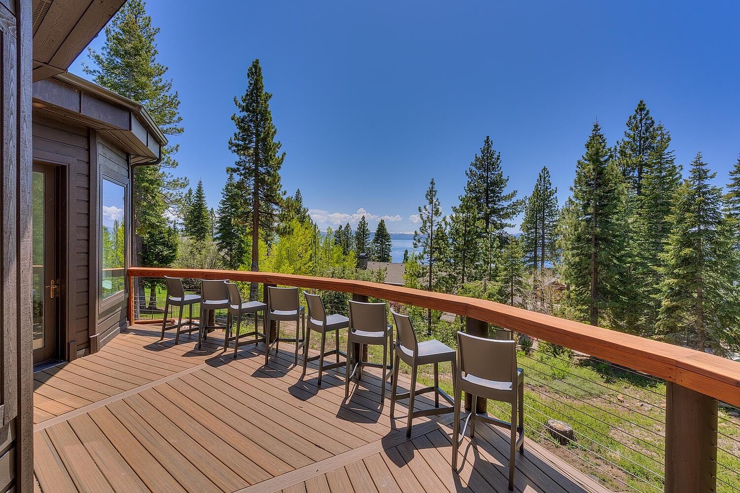 This image showcases a spacious wooden deck with a modern design, featuring a sleek railing and a row of stylish bar stools. The deck offers stunning views of the surrounding landscape, including lush trees and a glimpse of a serene lake under a clear blue sky. The scene evokes a sense of relaxation and outdoor living, perfect for entertaining or enjoying the natural beauty.