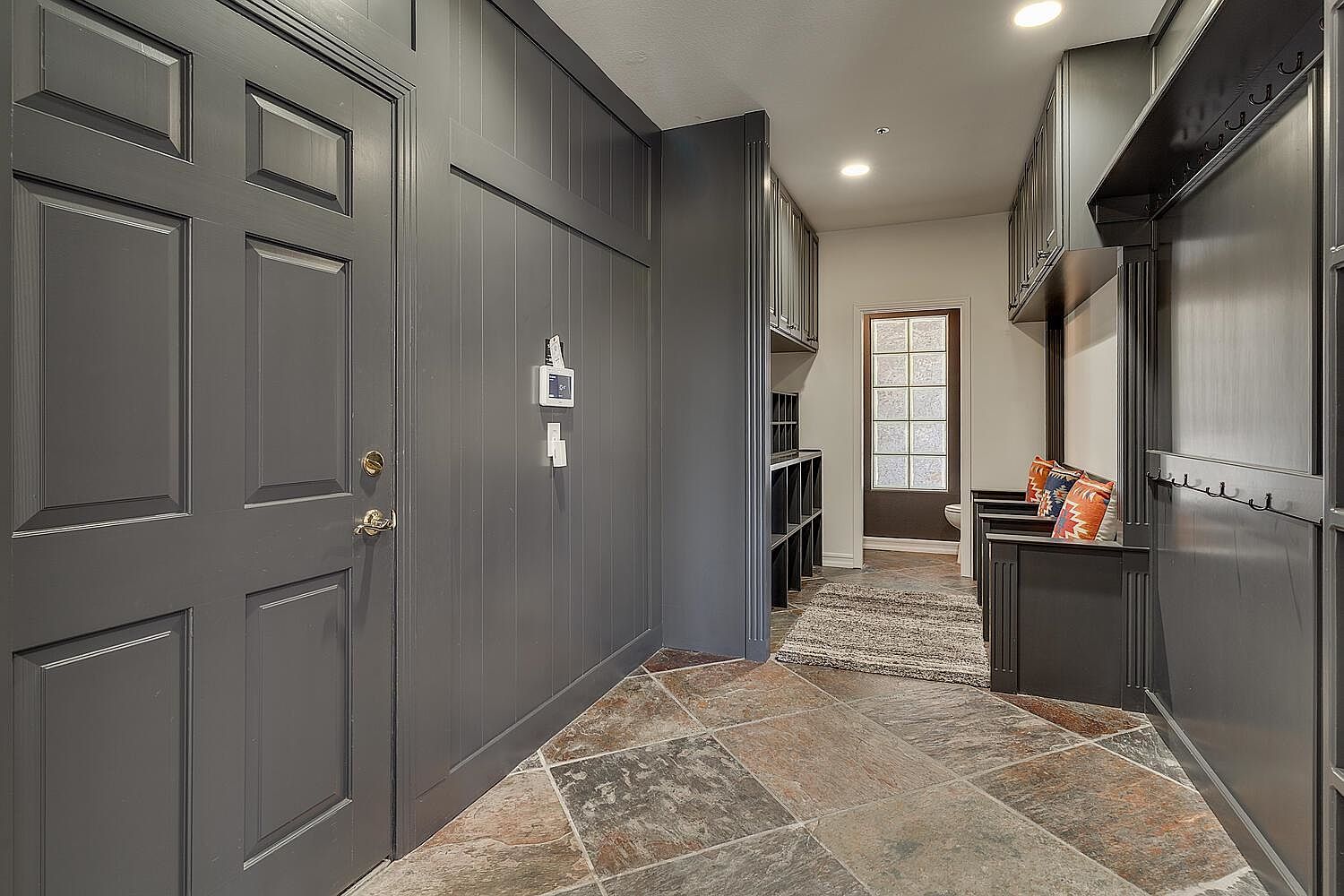 This image showcases a well-organized laundry room with dark gray cabinetry and a slate tile floor. The room features built-in storage, including cubbies and hooks, and a bench with decorative pillows. A window provides natural light, and the overall design is both functional and stylish.
