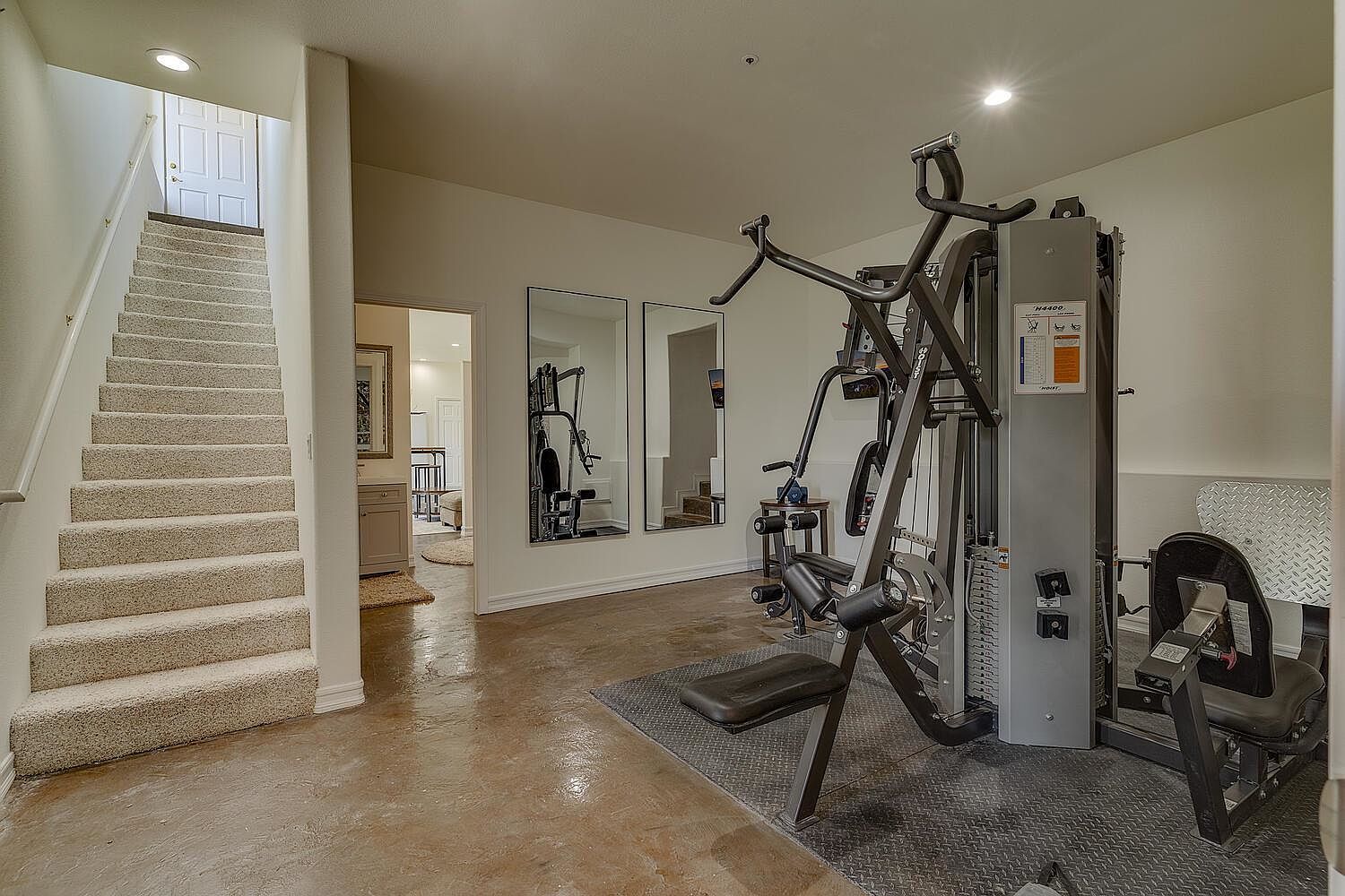 This is an interior shot of a home gym featuring a multi-station weight machine as the focal point. The room has polished concrete floors and two large mirrors on the wall, reflecting the equipment and space. A carpeted staircase leads to an upper level, and a doorway provides a glimpse into another room, suggesting a well-integrated and functional workout area.