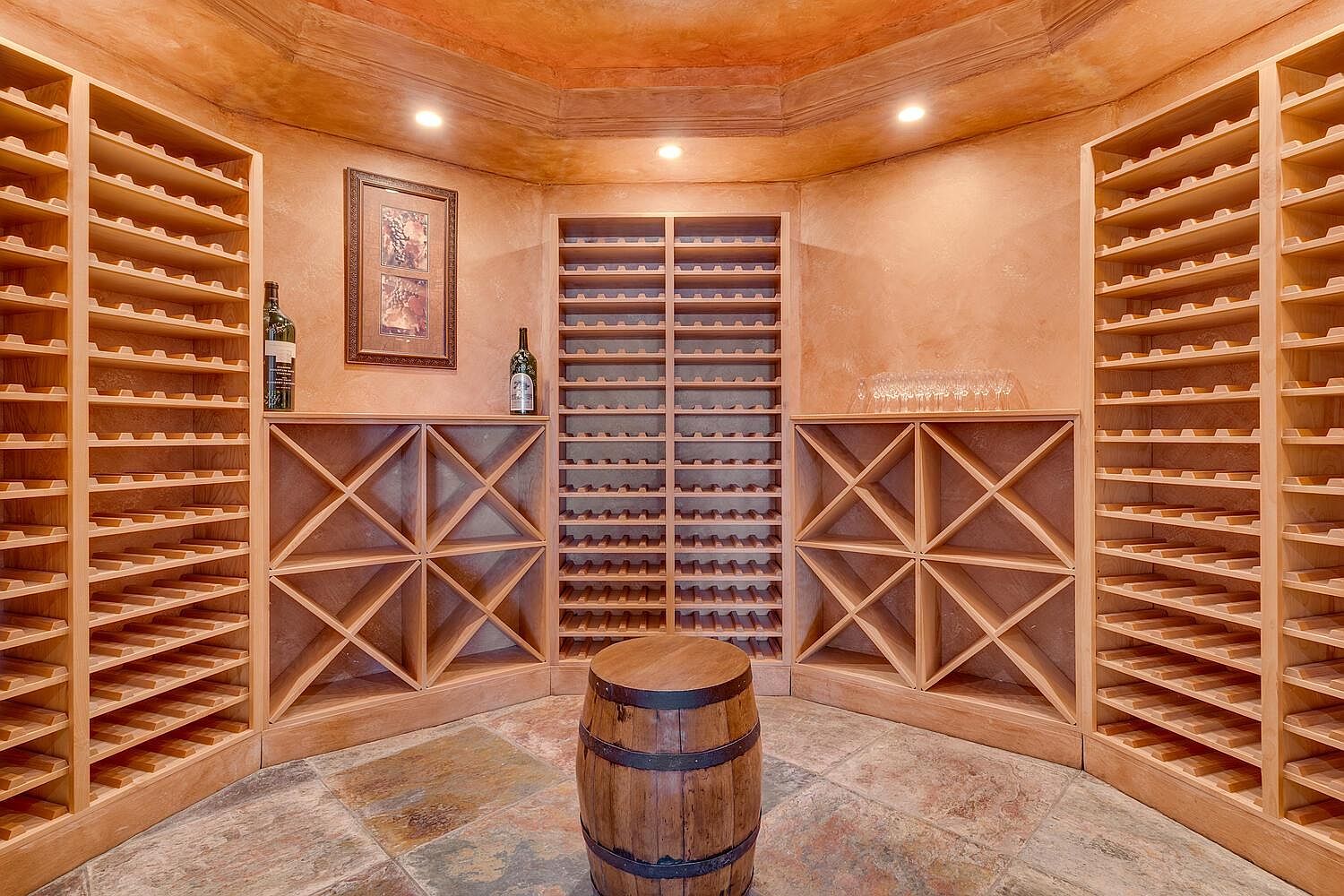 This is a well-organized wine cellar featuring wooden wine racks, a barrel in the center, and a stone floor. The walls are textured in a warm tone, and the lighting is soft, creating an inviting atmosphere. The cellar appears spacious and designed for wine storage and display.