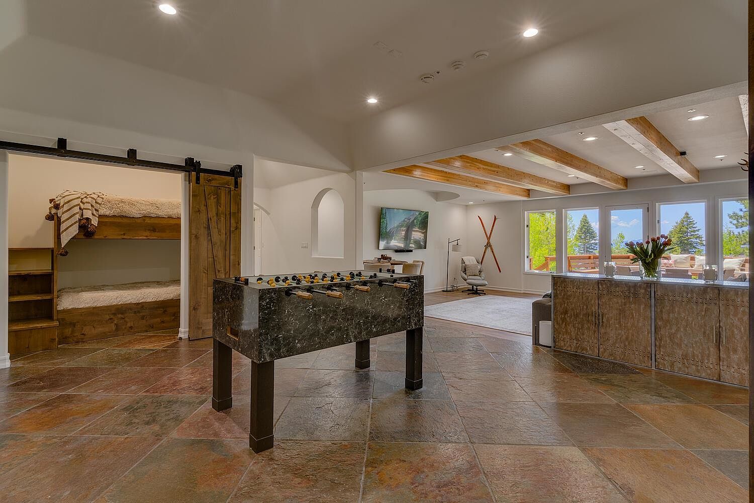 This is a spacious basement area featuring a foosball table as a central element. The room includes a bunk bed area with a sliding barn door, a living space with a TV, and a bar area with wooden cabinetry. The flooring is tiled with a multi-toned slate, and the overall style is rustic and inviting.