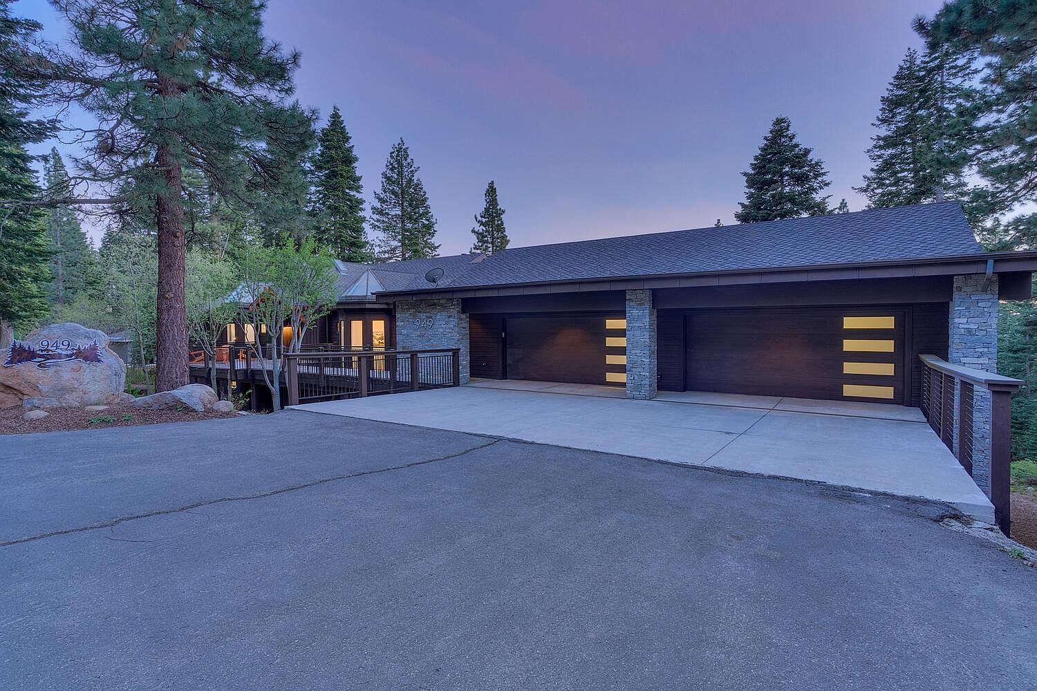 This is a front exterior view of a modern mountain home featuring a two-car garage with contemporary doors and stone accents. The driveway leads up to the garage and a deck area is visible to the left, surrounded by mature trees. The overall impression is one of luxury and privacy in a natural setting.